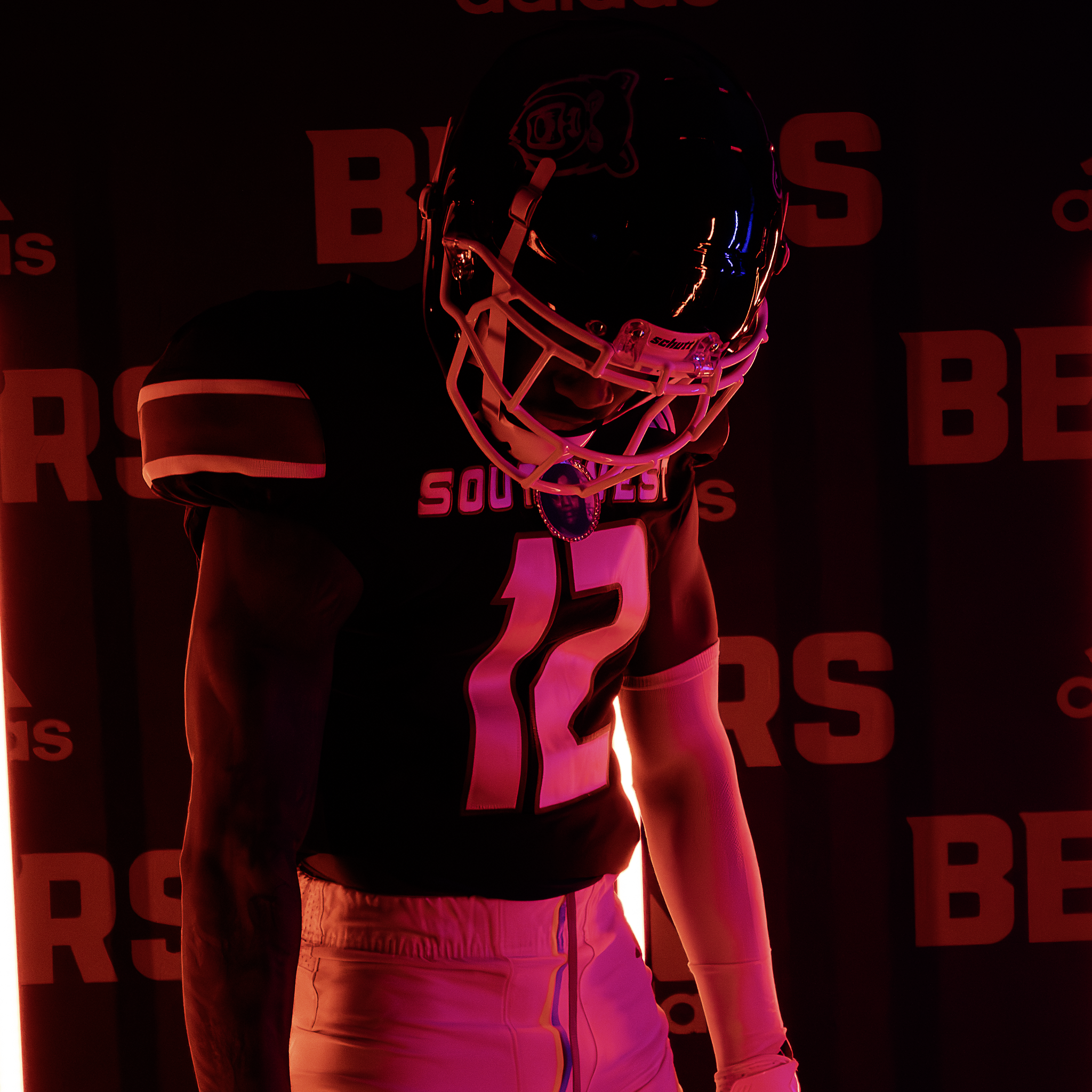 A football player wearing a black helmet with a lion logo, black jersey with white and red details, and white pants, standing in front of a backdrop with red and black text at a sports event.