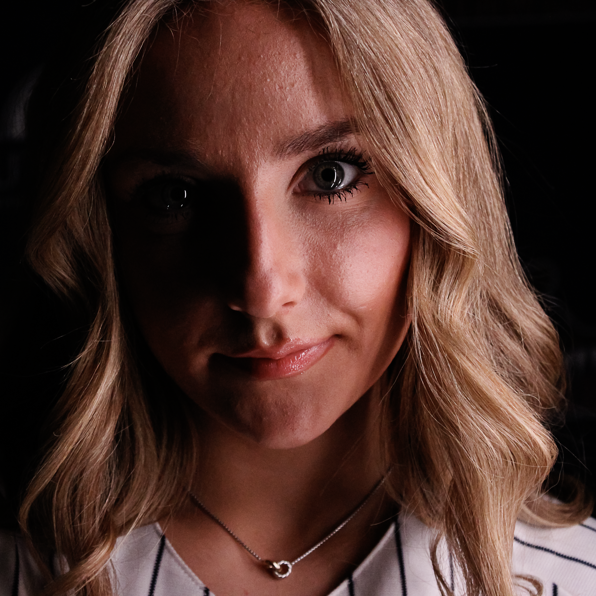 Close-up of a woman with blonde hair, blue eyes, and light skin, wearing a silver necklace with a heart-shaped pendant and a white top with black stripes. She has makeup with dark eyeliner and mascara.