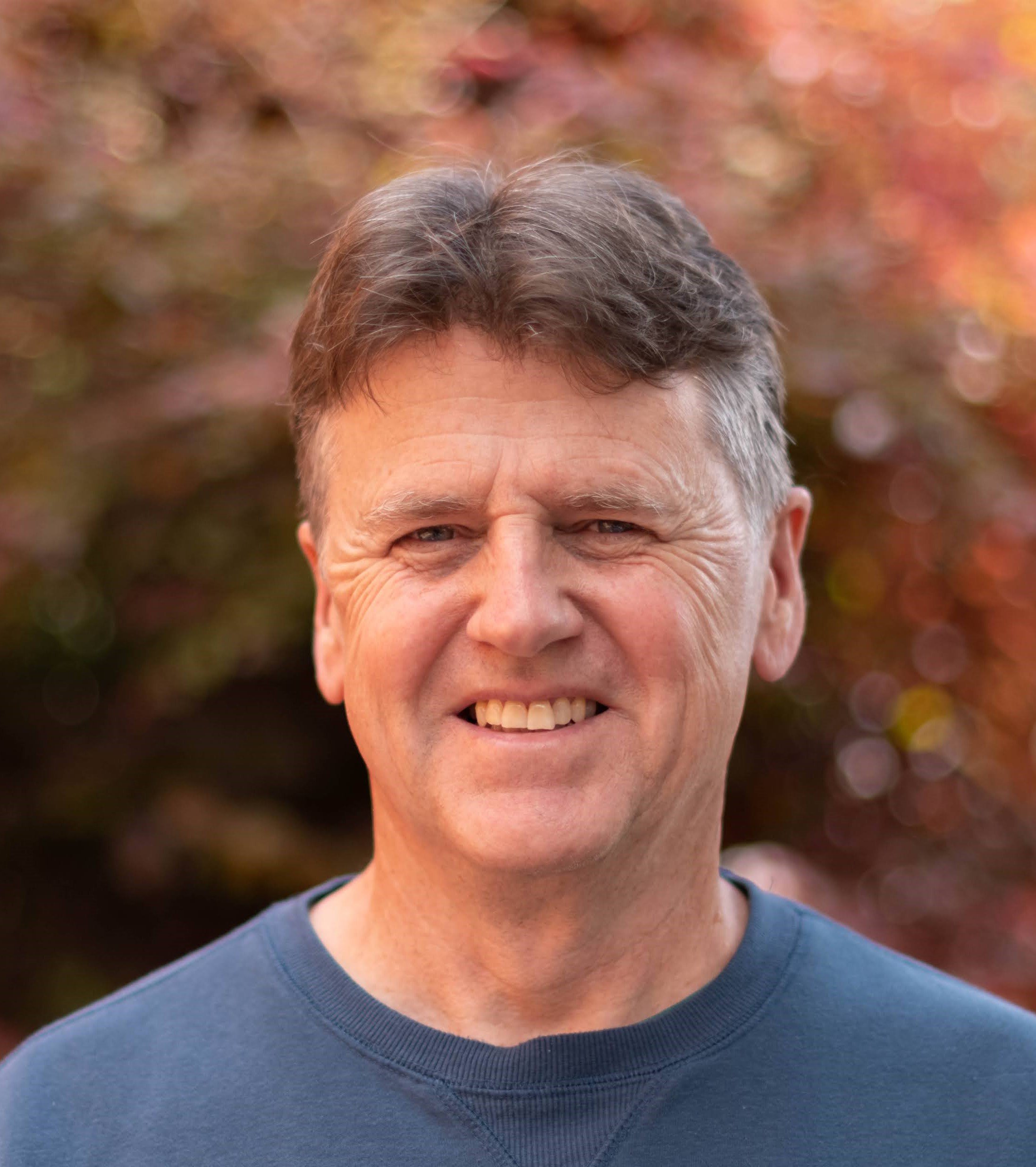 Close-up of a smiling middle-aged man with short, gray hair, wearing a gray crew-neck shirt, outdoors with blurred autumn-colored foliage in the background.
