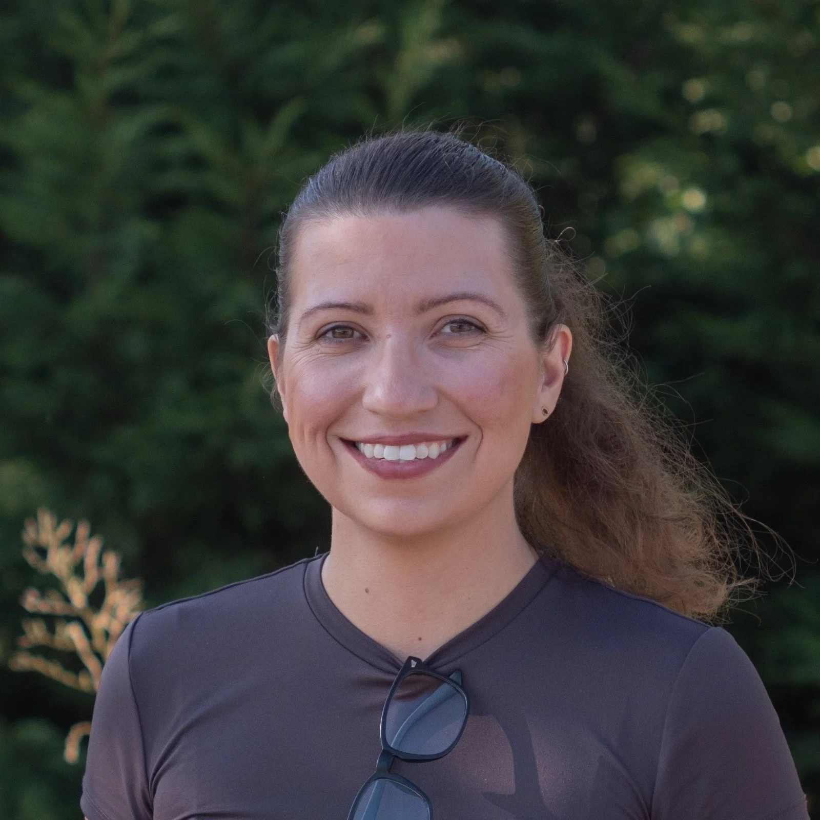 A woman with brown hair tied back, smiling, standing outdoors with green trees in the background, wearing a dark gray athletic shirt with sunglasses hanging from her shirt.