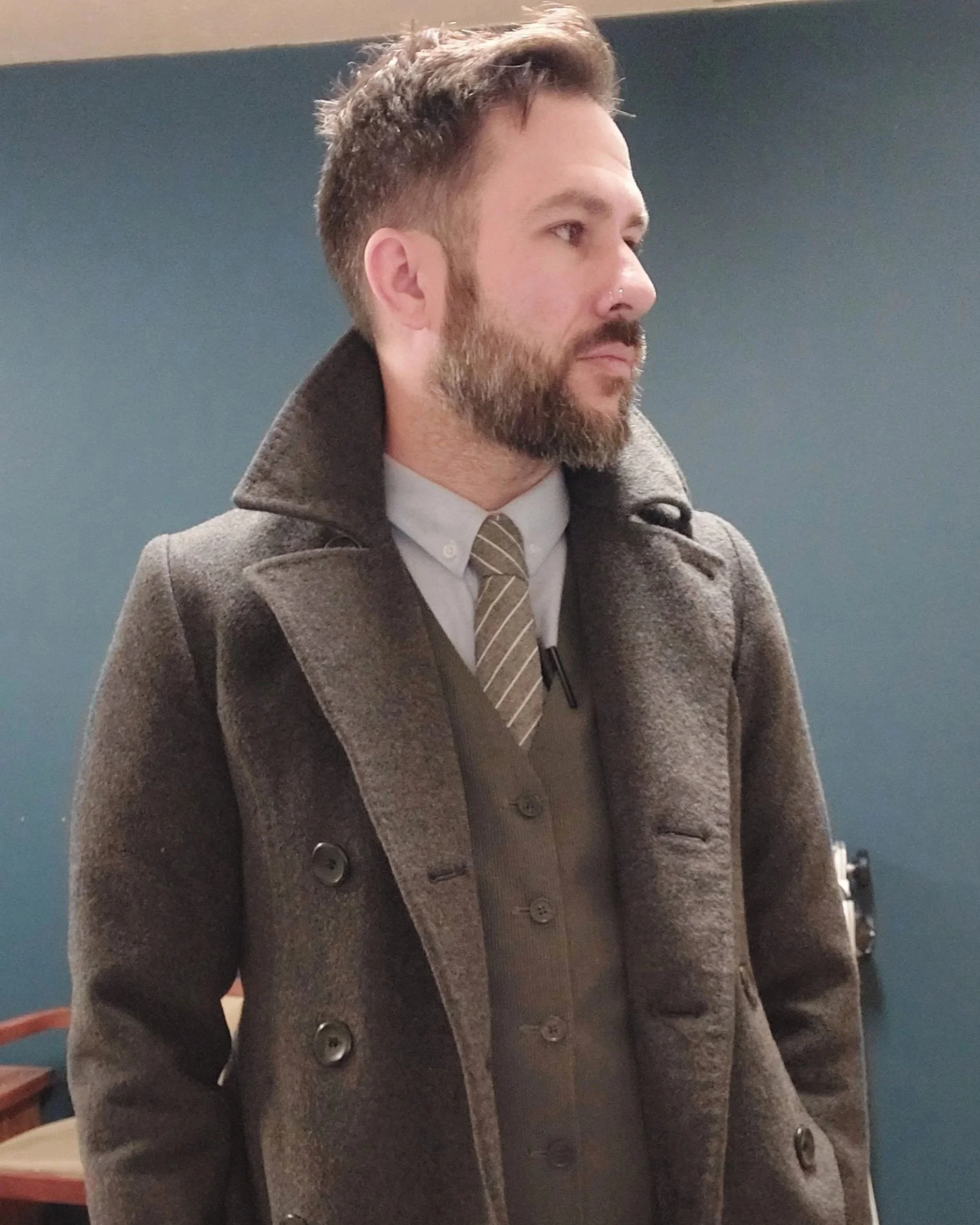 A man with a beard and styled hair wearing a gray coat, white shirt, and striped tie, standing indoors against a teal wall.