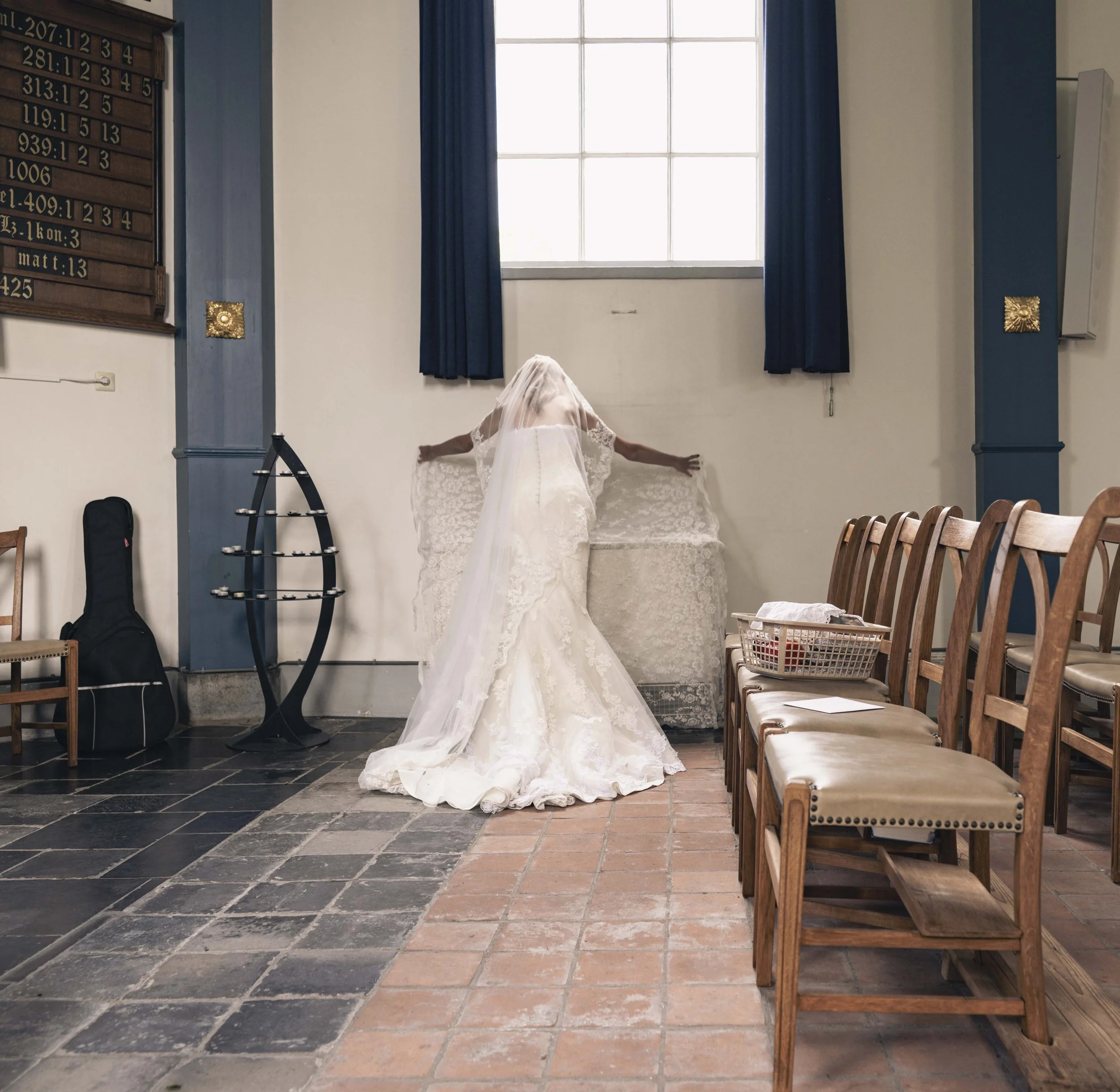 Binnenruimte van een kerk met houten banken, een groot raam met blauwe gordijnen en een bruid die zich klaar maakt.