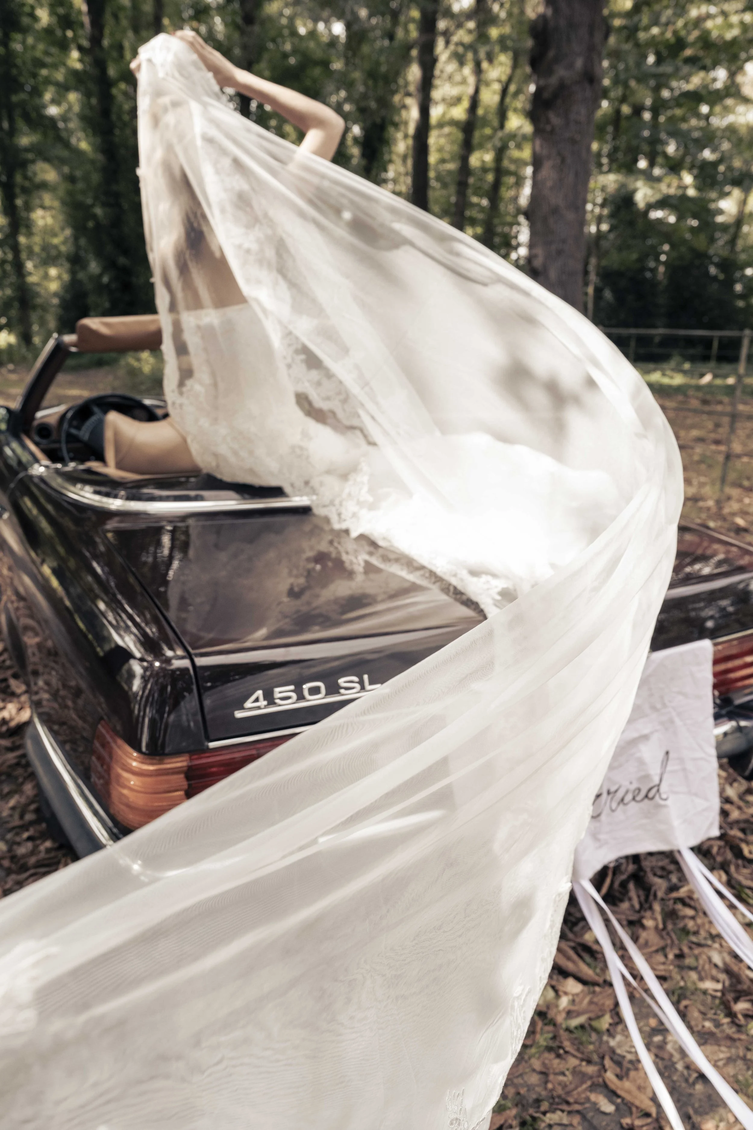 Een automodel, een Mercedes 450 SL, wordt gedecoreerd voor een bruiloft met een witte sluier en bloemen, omringd door bomen in een bosachtige omgeving.