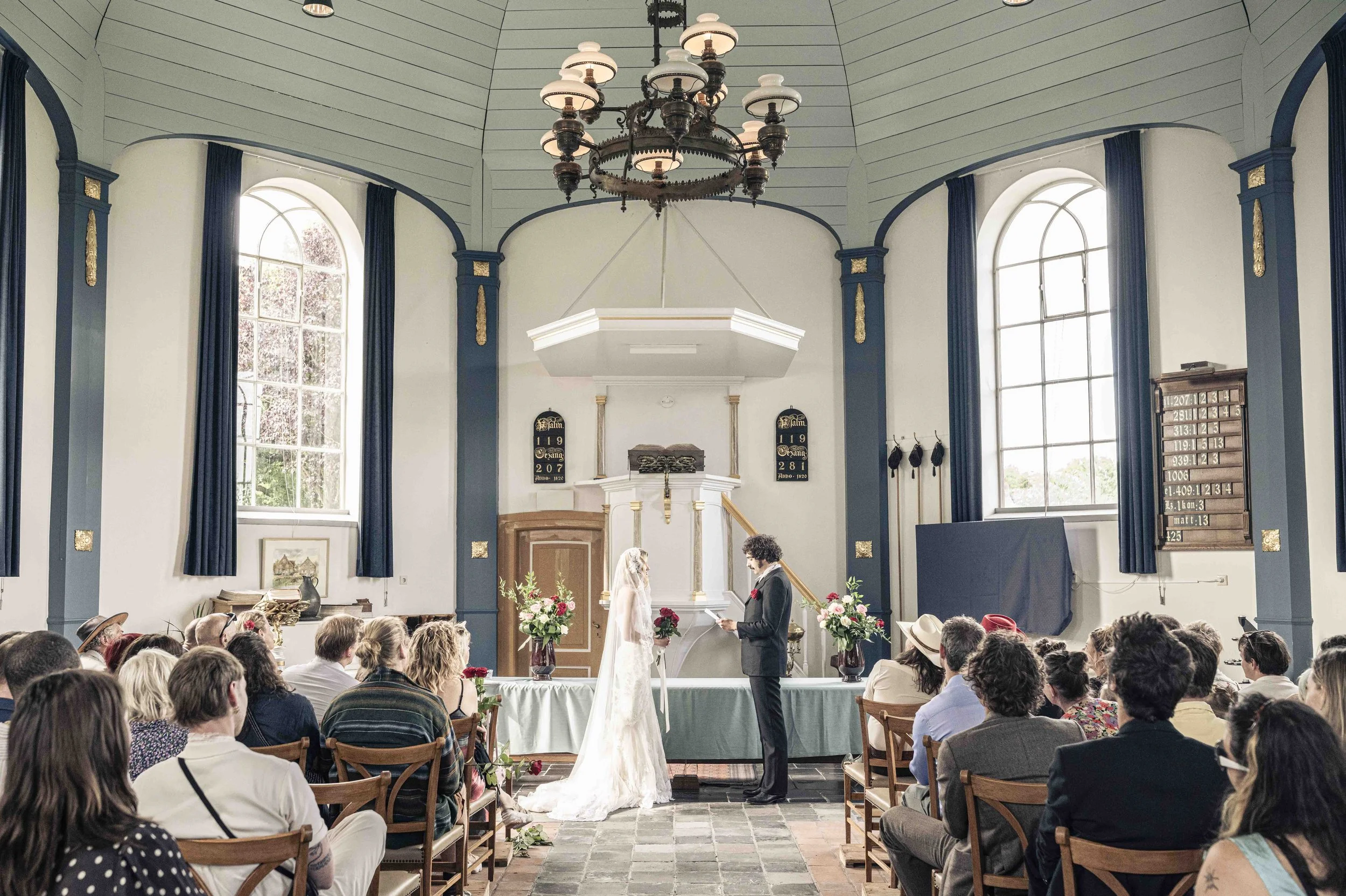 Trouwplechtigheid in een mooie kerk met hoge ramen, een kroonluchter en een publiek van gasten die naar het bruidspaar kijken.