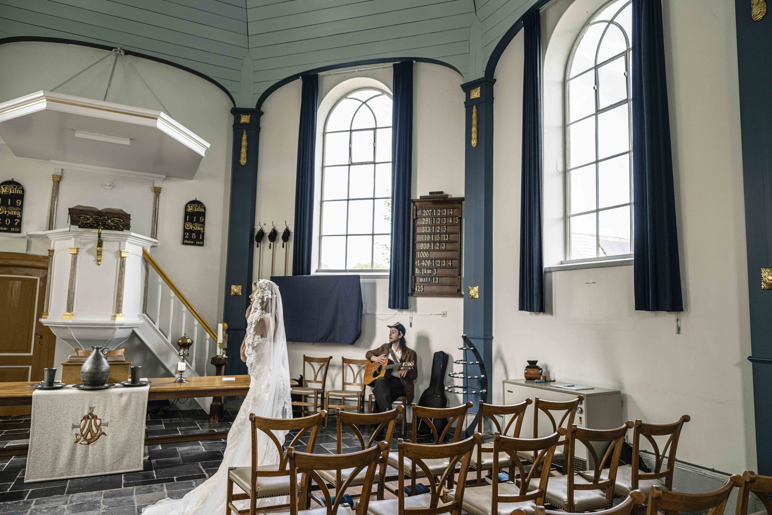 Binnenkant van een kerk met grote ramen en blauwe gordijnen, enkele mensen, waaronder een vrouw in een witte trouwjurk en een man met een gitaar, mogelijk tijdens een huwelijksceremonie.