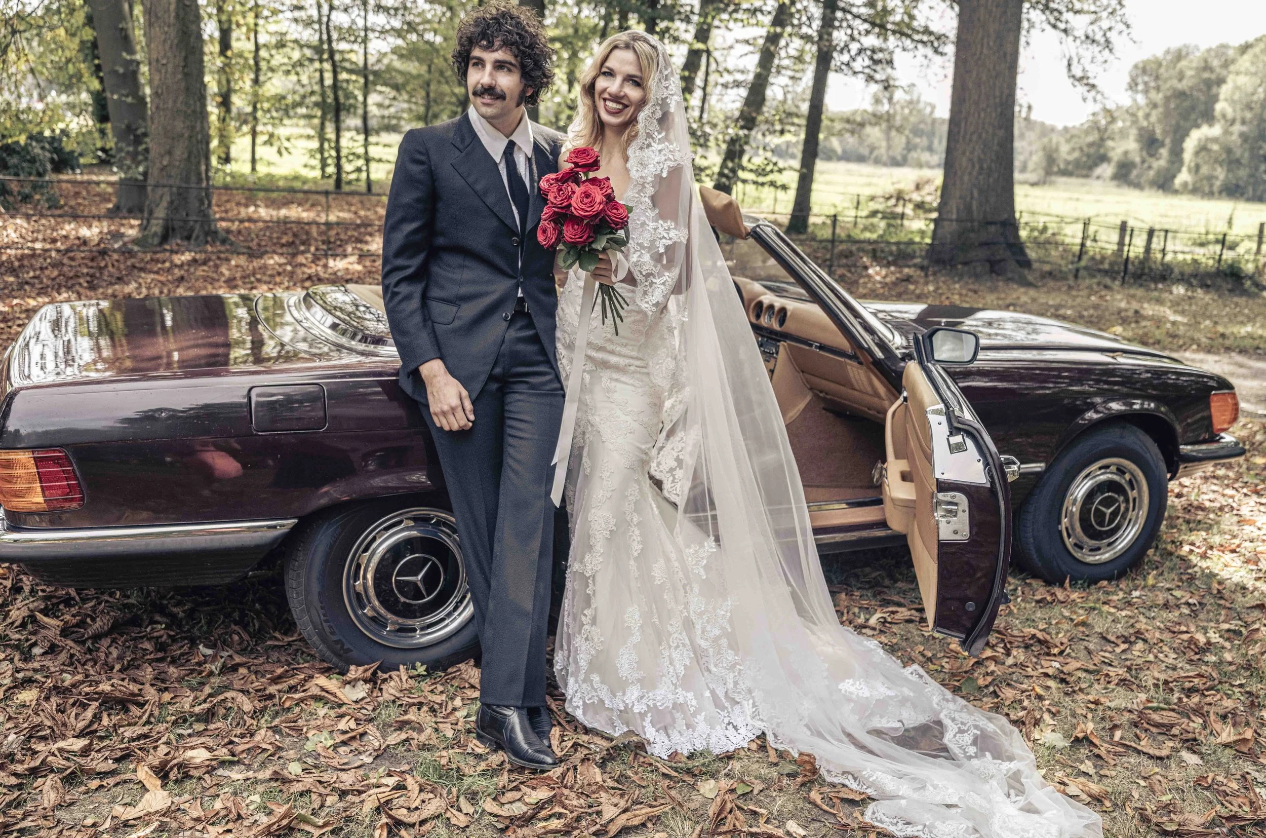 Een getrouwd stel poseert voor een zwarte Mercedes-Benz Cabriolet in een bosrijke omgeving, met herfstbladeren op de grond. De bruid draagt een witte kanten trouwjurk met lange sluier en houdt een boeket roderozen vast. De bruidegom draagt een donker