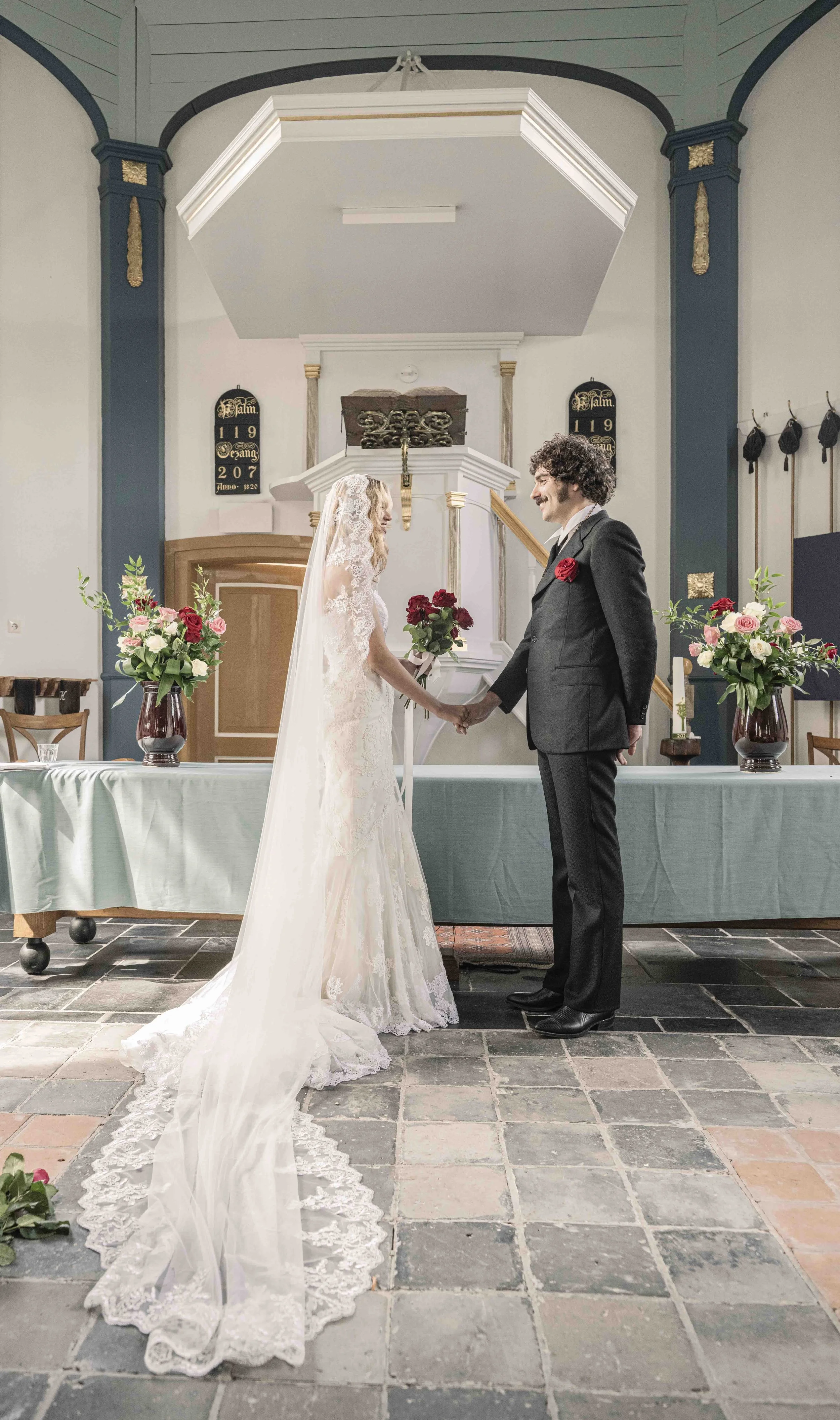 Een bruid en bruidegom houden elkaars handen vast tijdens een huwelijk in een kerk, omringd door bloemen en traditionele decoraties.