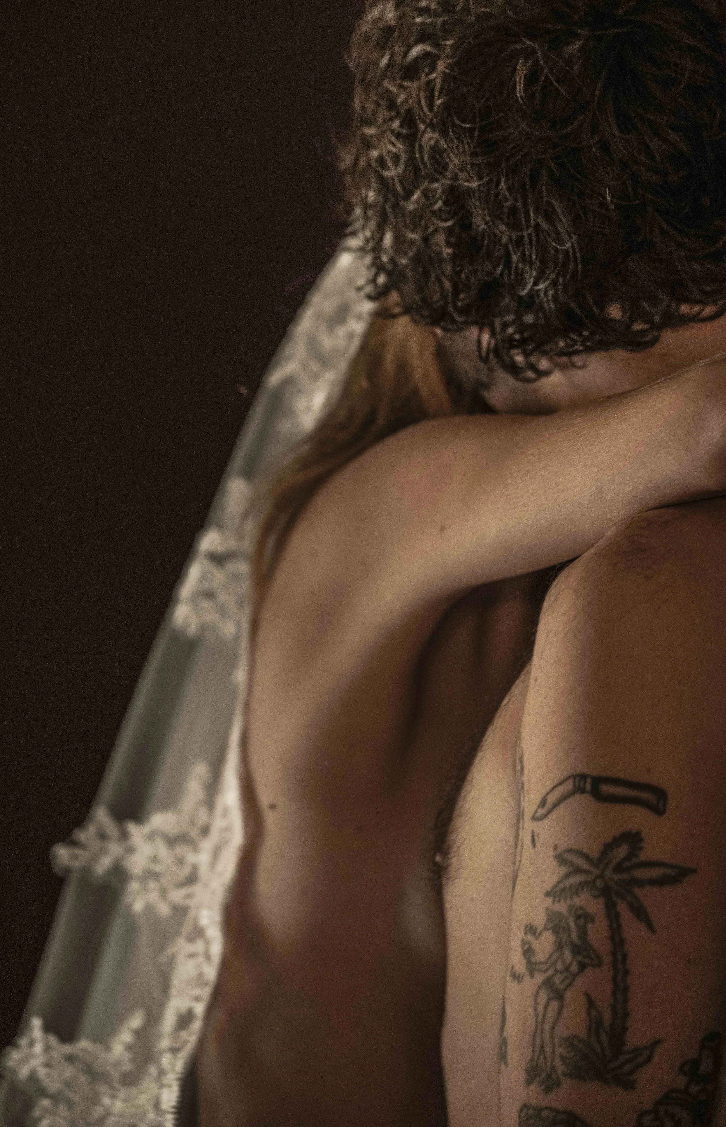 Close-up van een persoon met getatoeëerde arm, die een stuk stof of kleding omhoog houdt, met een achtergrond van donkere ruimte en een stuk kantachtige stof.