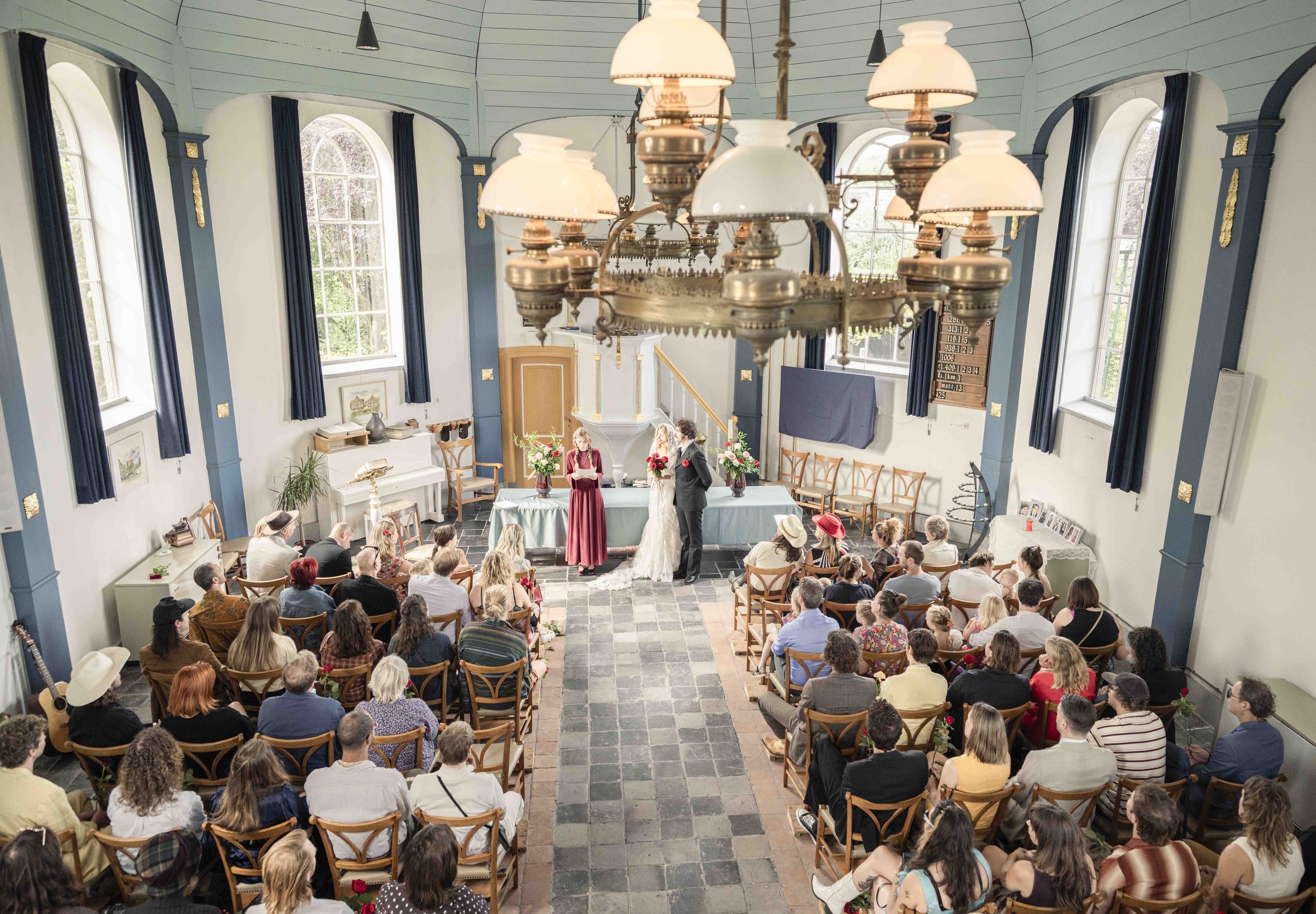 Een kerkelijke ceremonie met een bruid en bruidegom voor een publiek. Mensen zitten in houten stoelen, sommige dragen hoedjes. De kerk is lichtblauw met hoge ramen en een grote kroonluchter hangt aan het plafond.