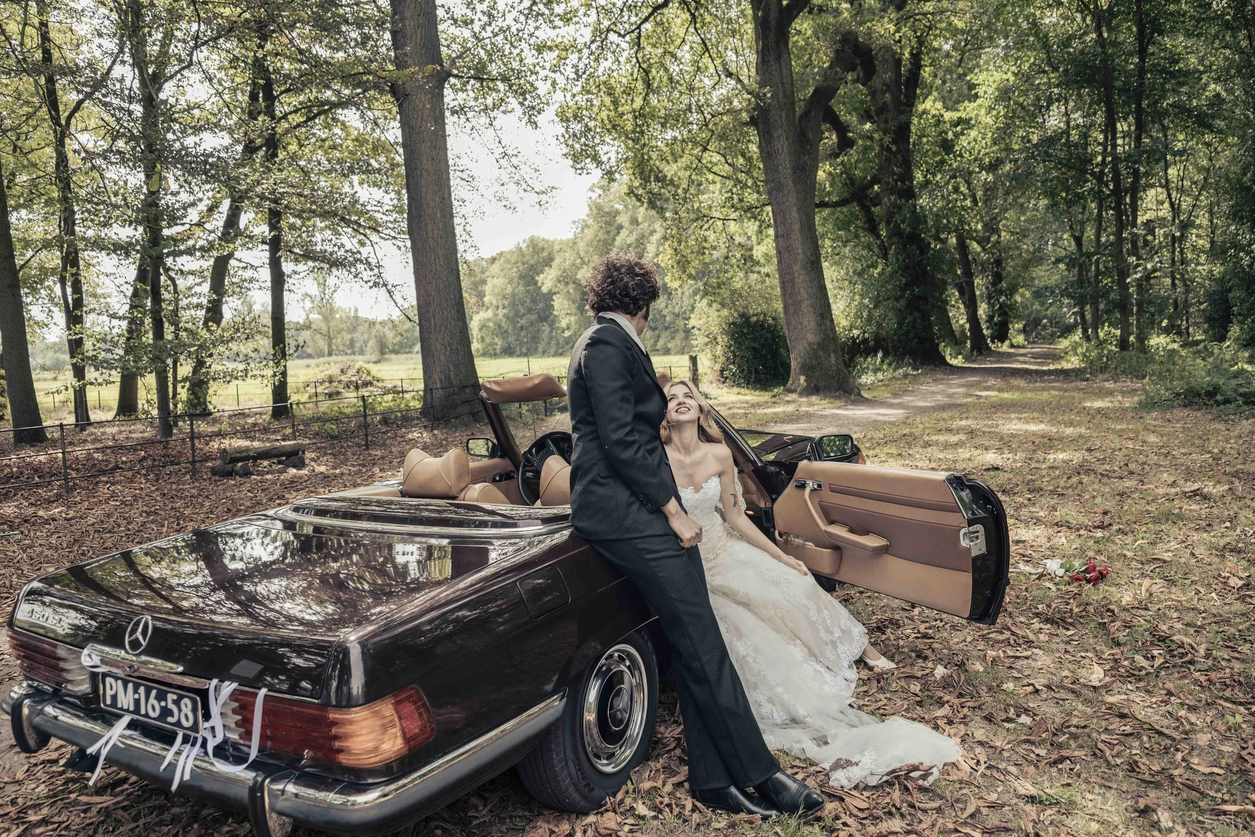 Een bruid en een bruidegom staan naast een zwarte Mercedes-Benz cabriolet in een bosrijke omgeving. De bruid zit in de auto met haar voeten buiten de deur, terwijl de bruidegom naast haar staat, samen in een bos met bladeren op de grond en hoge bomen