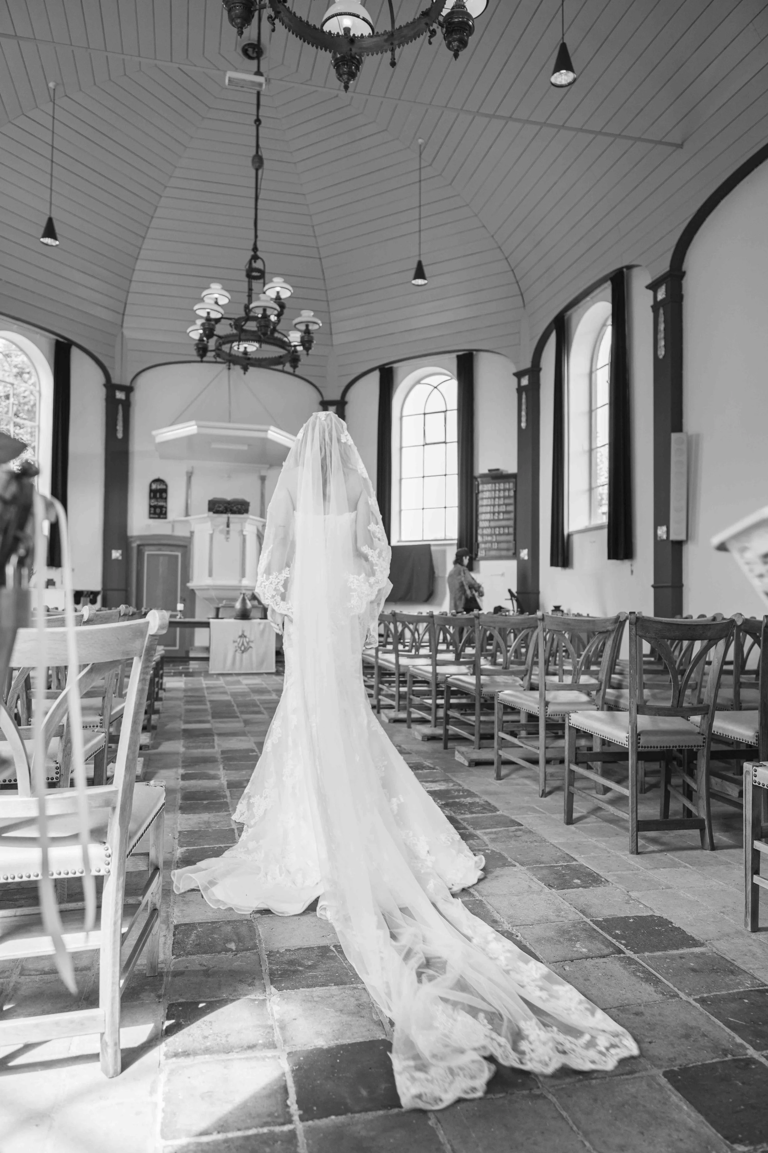 Een binnenzijde van een kerk of kapel, met rijen houten stoelen, een kroonluchter aan het plafond, en een witte trouwjurk met lange sleep en kanten details hangend in het midden van de ruimte.