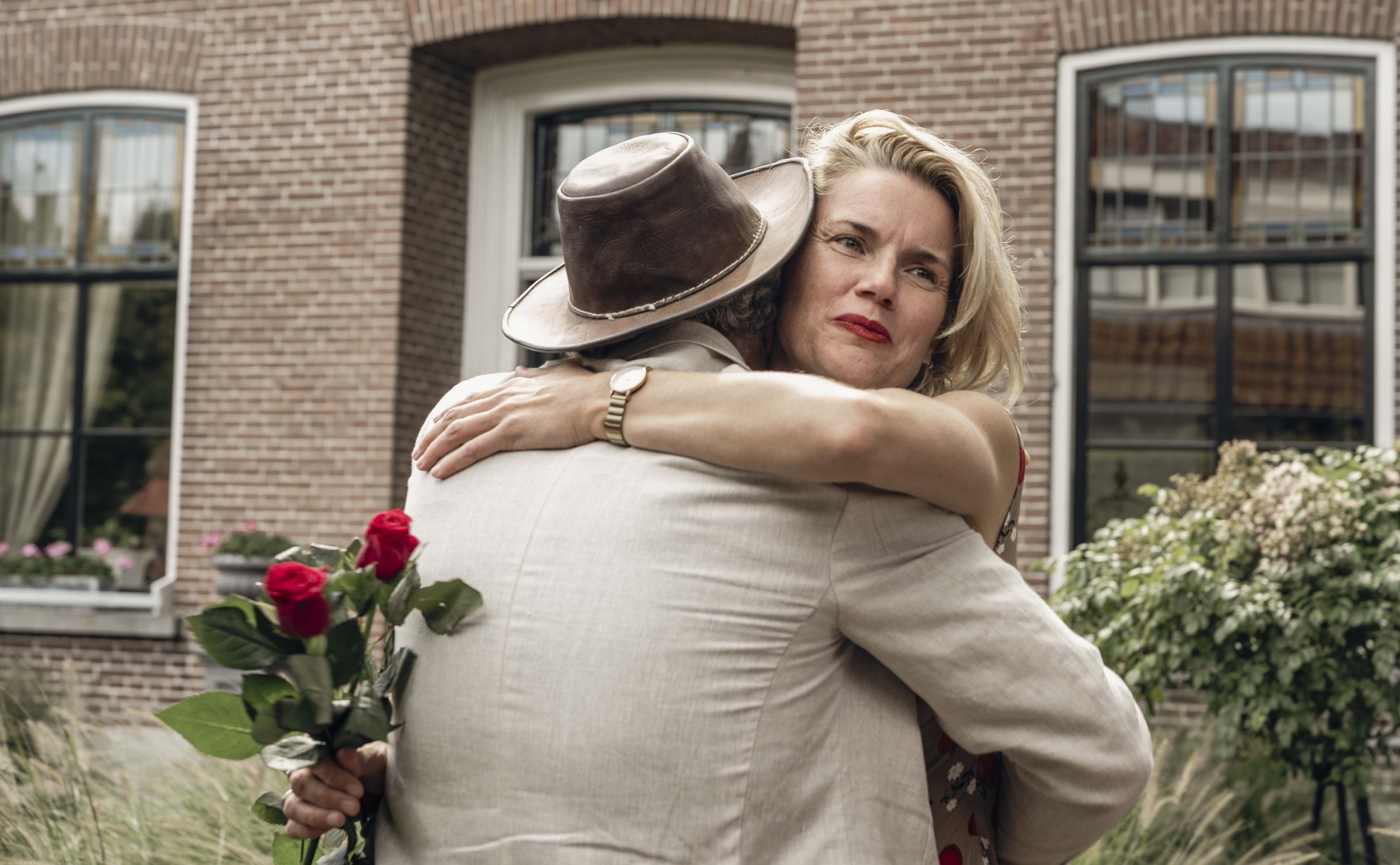 Een vrouw wordt door een man omarmd terwijl ze een bos rode rozen vasthoudt, buiten voor een bakstenen huis.