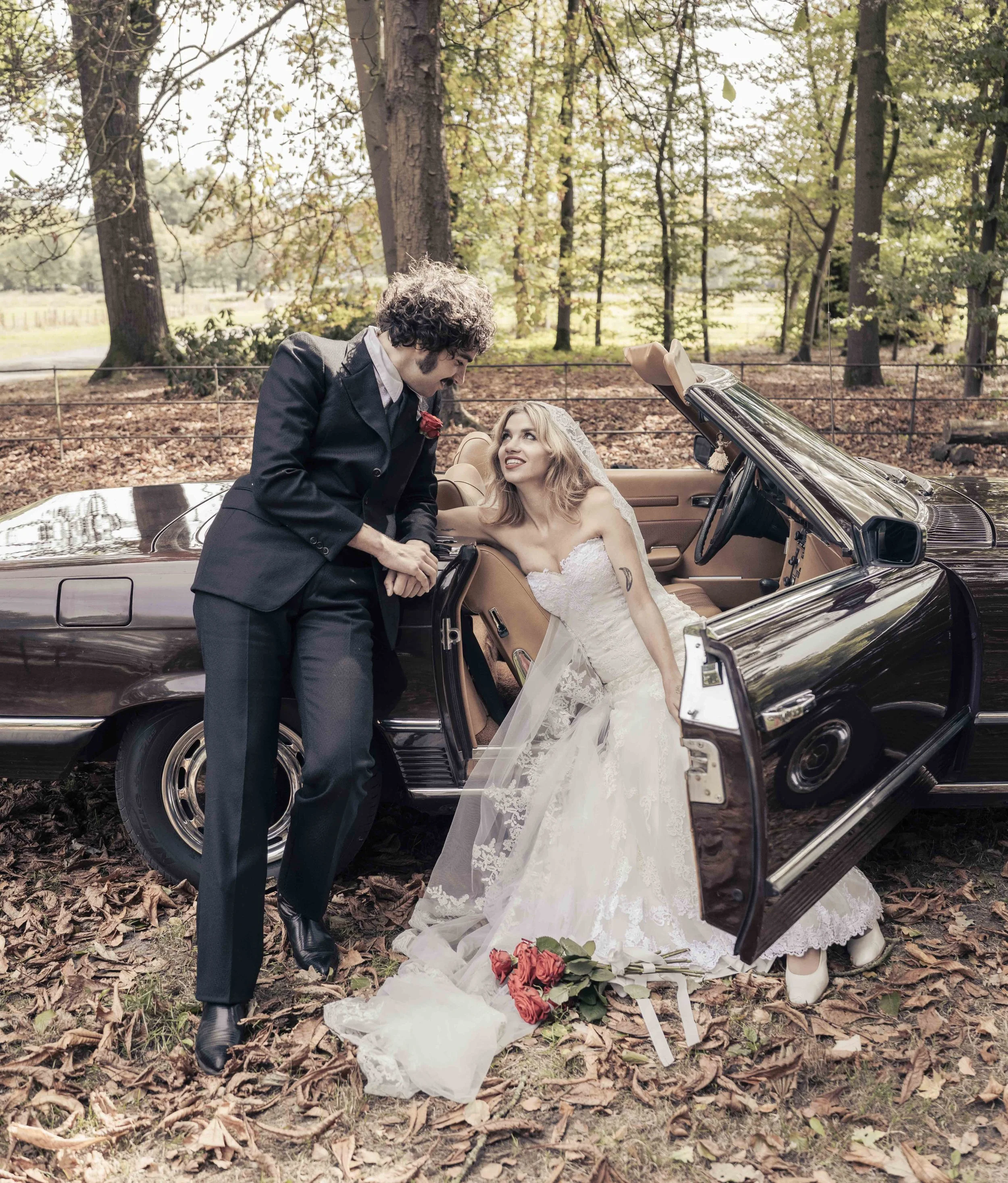 Een man en een vrouw die op een bosgrond staan, de man knielt en lijkt de vrouw te knuffelen of verzorgen. De vrouw ligt in een open autocabine in een bruin cabriolet en kijkt omhoog. De vrouw draagt een witte jurk en een hoofdband. Er liggen bloemen