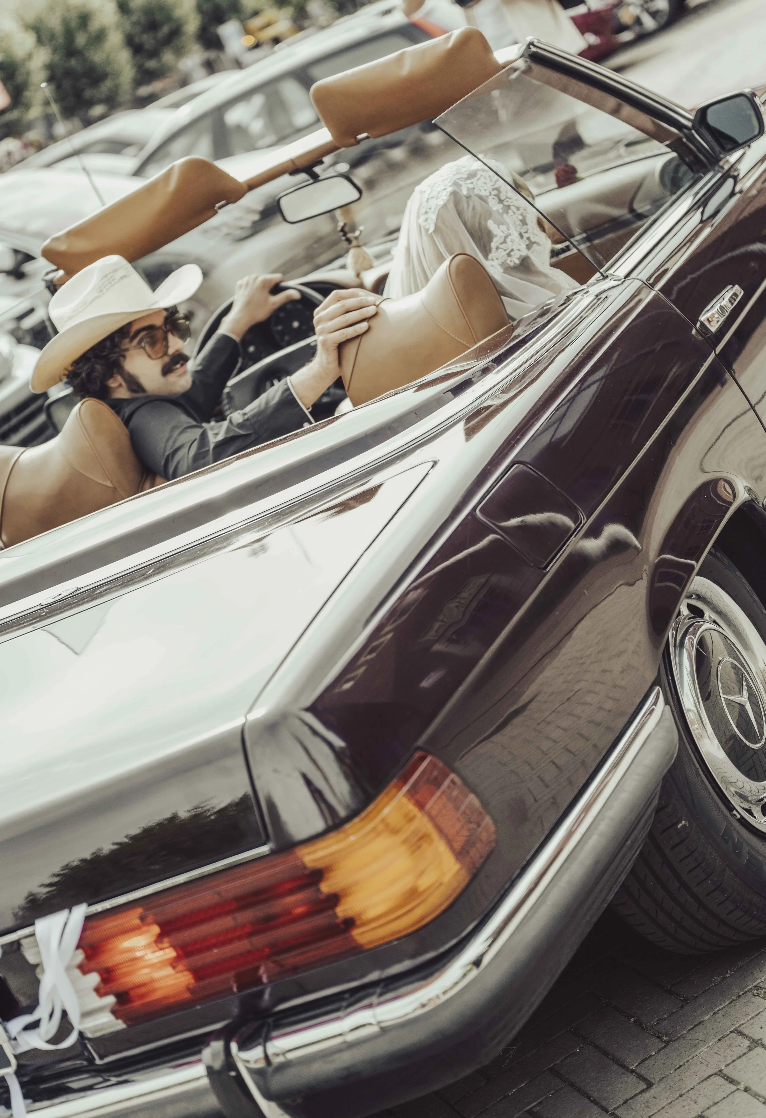 Een persoon rijdt in een klassieke zwarte cabriolet met leren interieur, gekleed in een zwart pak en draagt een cowboyhoed en een brillen. De persoon houdt de stuur en is te zien door de voorruit.