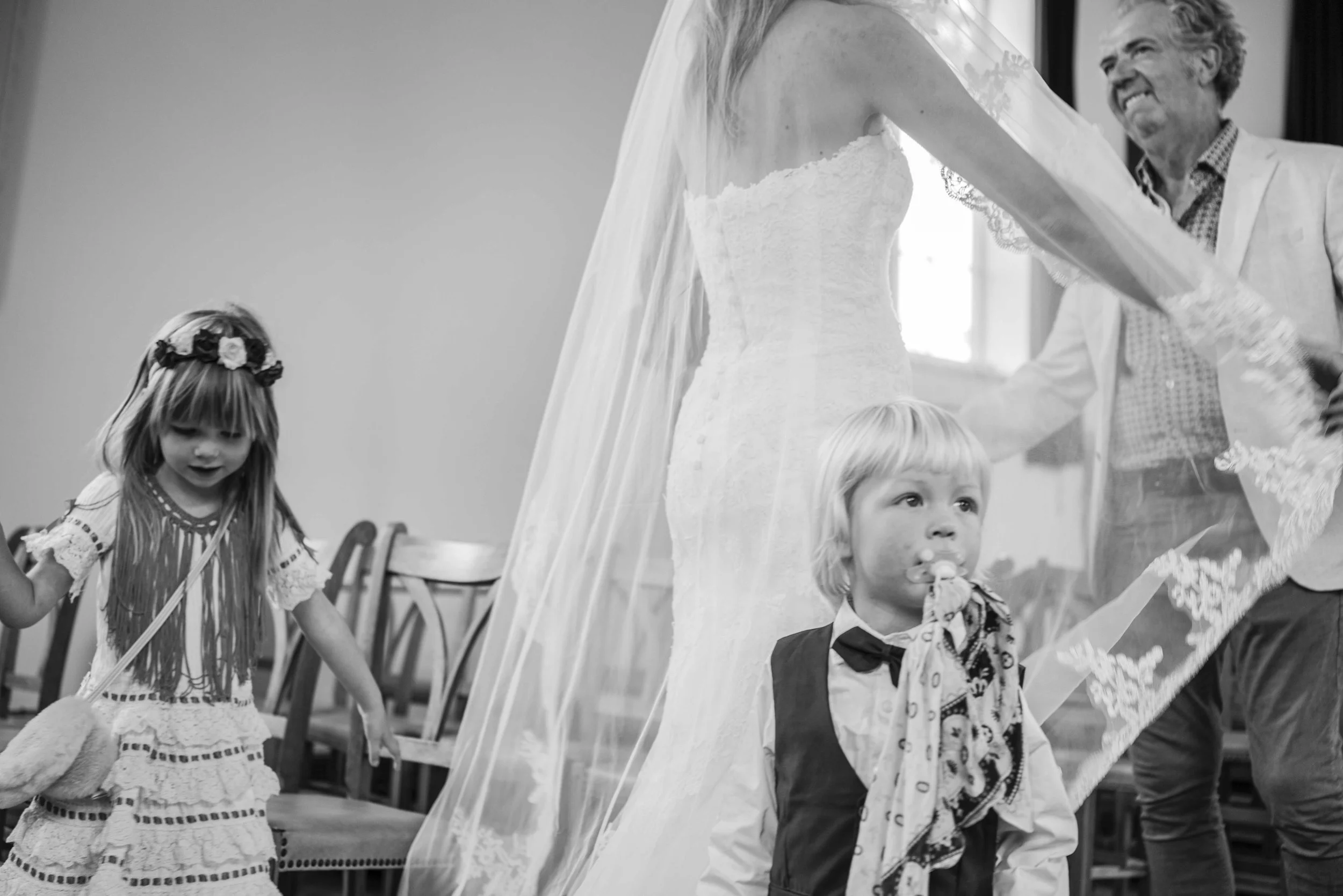 Twee jonge kinderen, een meisje en een jongen, met een volwassen vrouw in witte jurken en de man in een lichte jas, binnen een ruimte met stoelen.