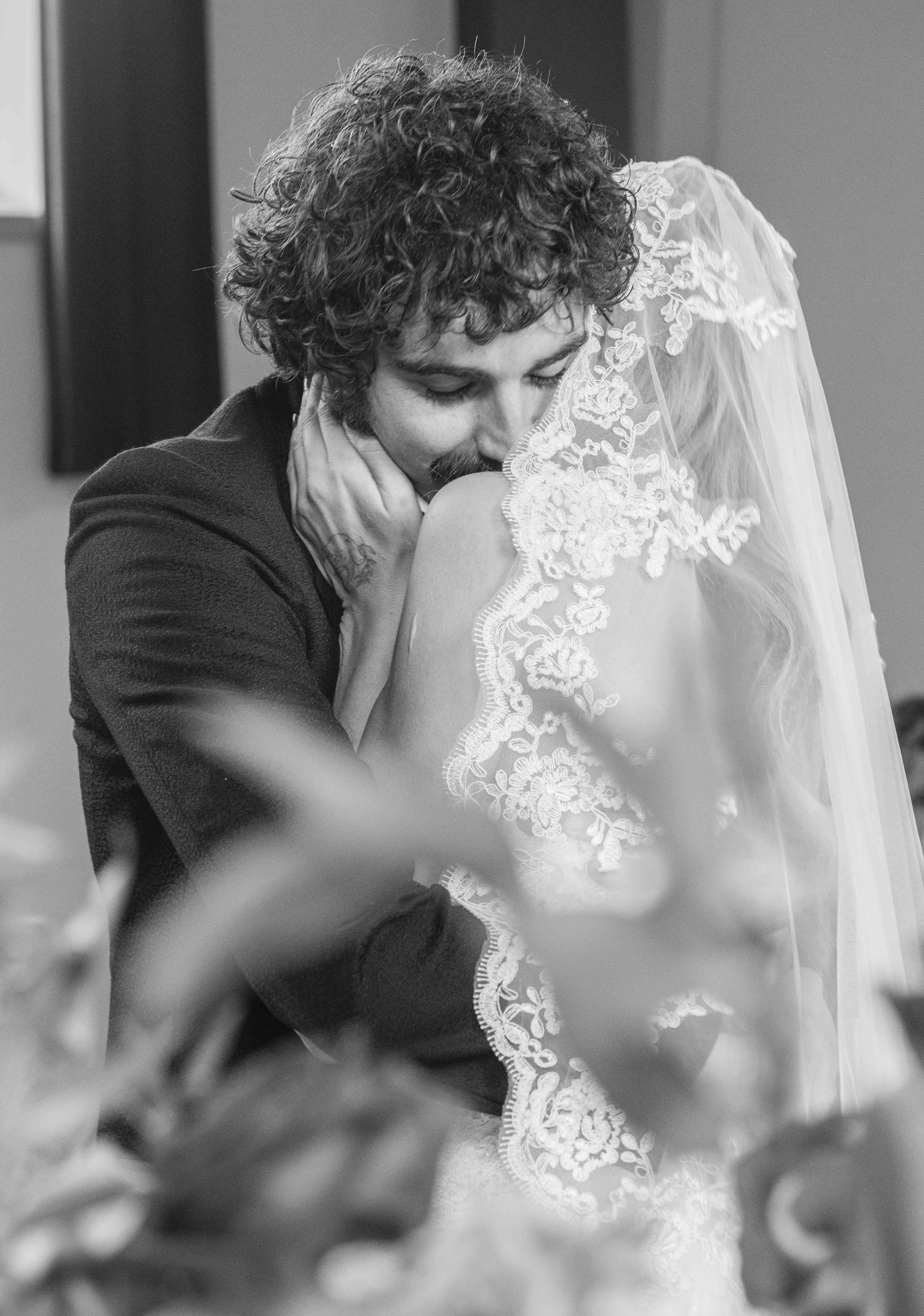 In de afbeelding wordt een emotionele scène weergegeven van een persoon die een ander omhelst, mogelijk tijdens een bruiloft. Het is een zwart-wit foto en de persoon die omhelst draagt een kanten trouwjurk en een sluier.