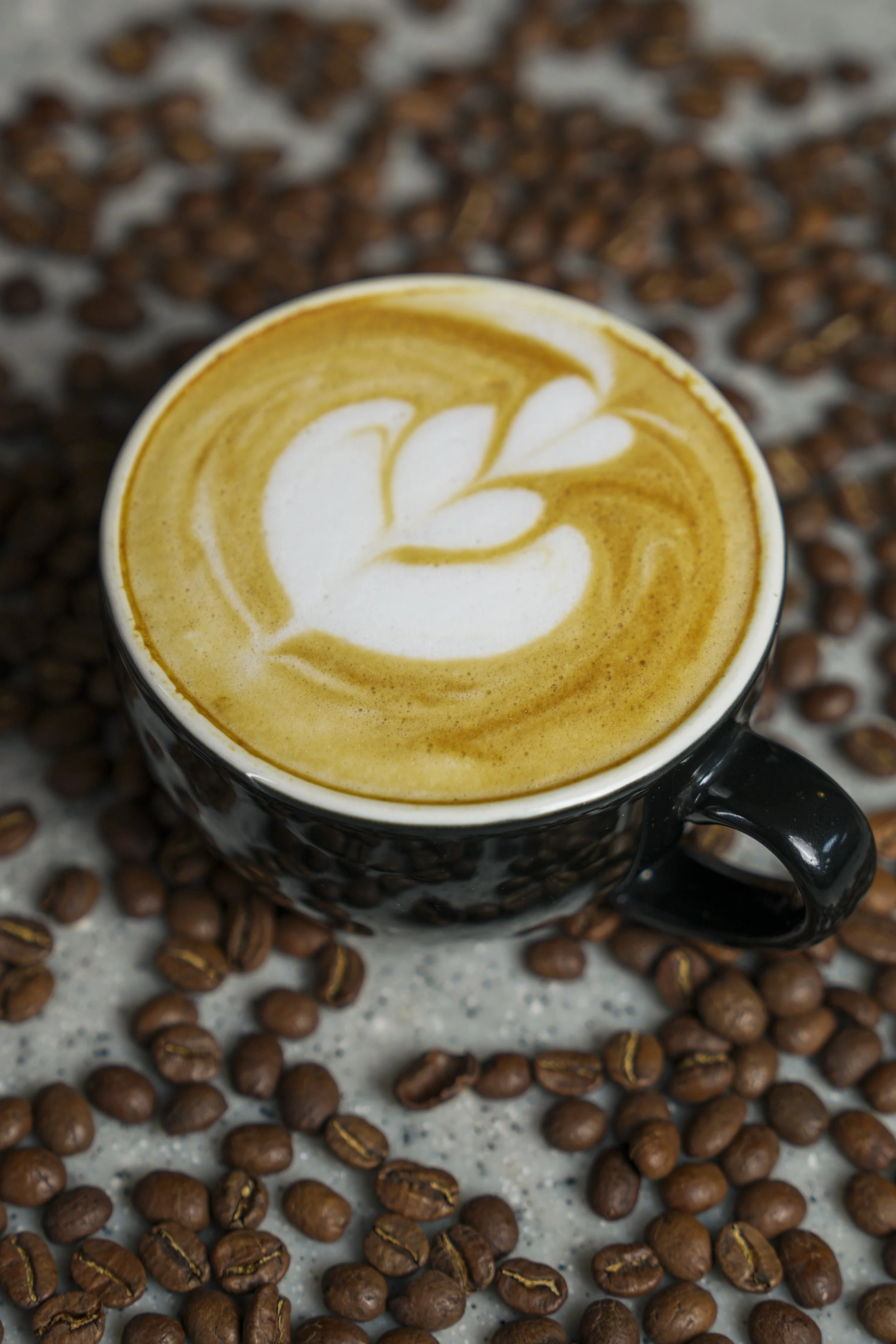 A black ceramic cup filled with a latte displaying latte art in the shape of a leaf, surrounded by scattered coffee beans on a grey surface.