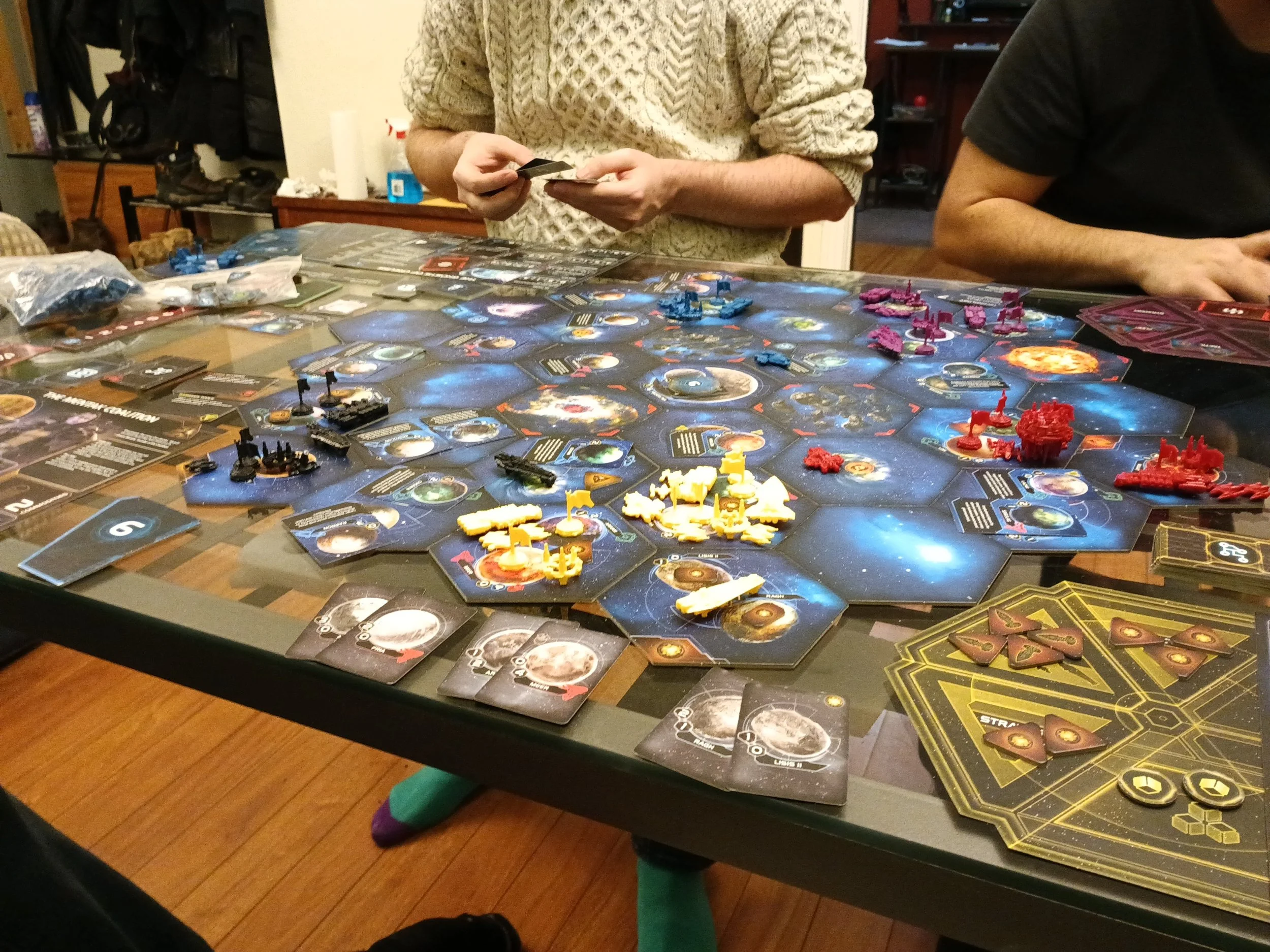 Jeu de société spatial (Twilight Imperium) avec des cartes, des pièces colorées et des figurines, des deux côtés d'une table entourée d'amis en train de jouer avec le coaching d'un animateur de Tables Rondes.