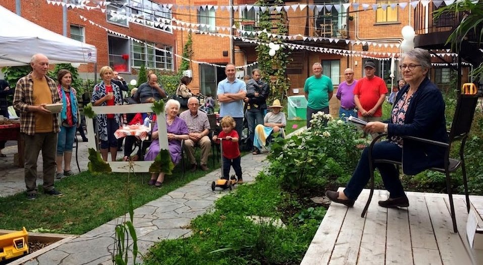 Un groupe de personnes, y compris des enfants et des personnes âgées, assistent à une assemblée spéciales pour l'anniversaire d'une coopérative d'habitation. Une animatrice est assise sur une plateforme tandis que les autres écoutent dans le jardin.