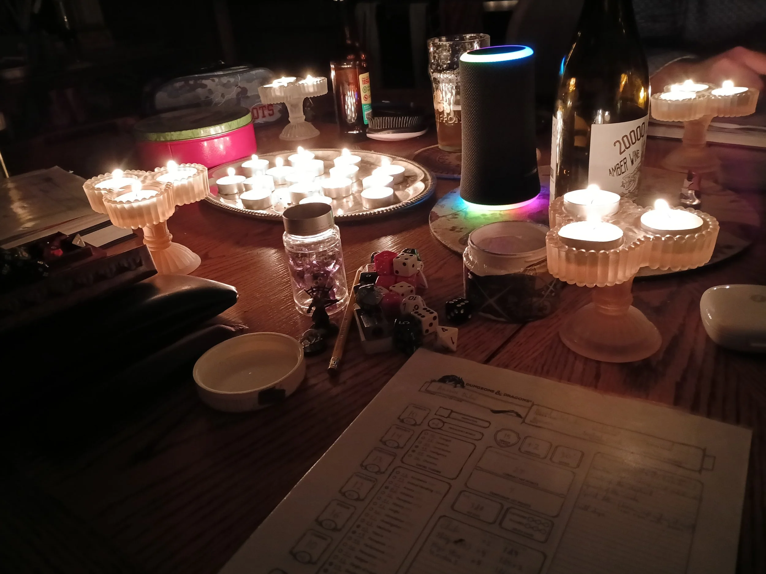 Une table avec plusieurs bougies allumées, un haut-parleur intelligent avec une lumière colorée, diverses boissons, dés à jouer, une assiette en argent, et un jeu de société avec des feuilles de règles.