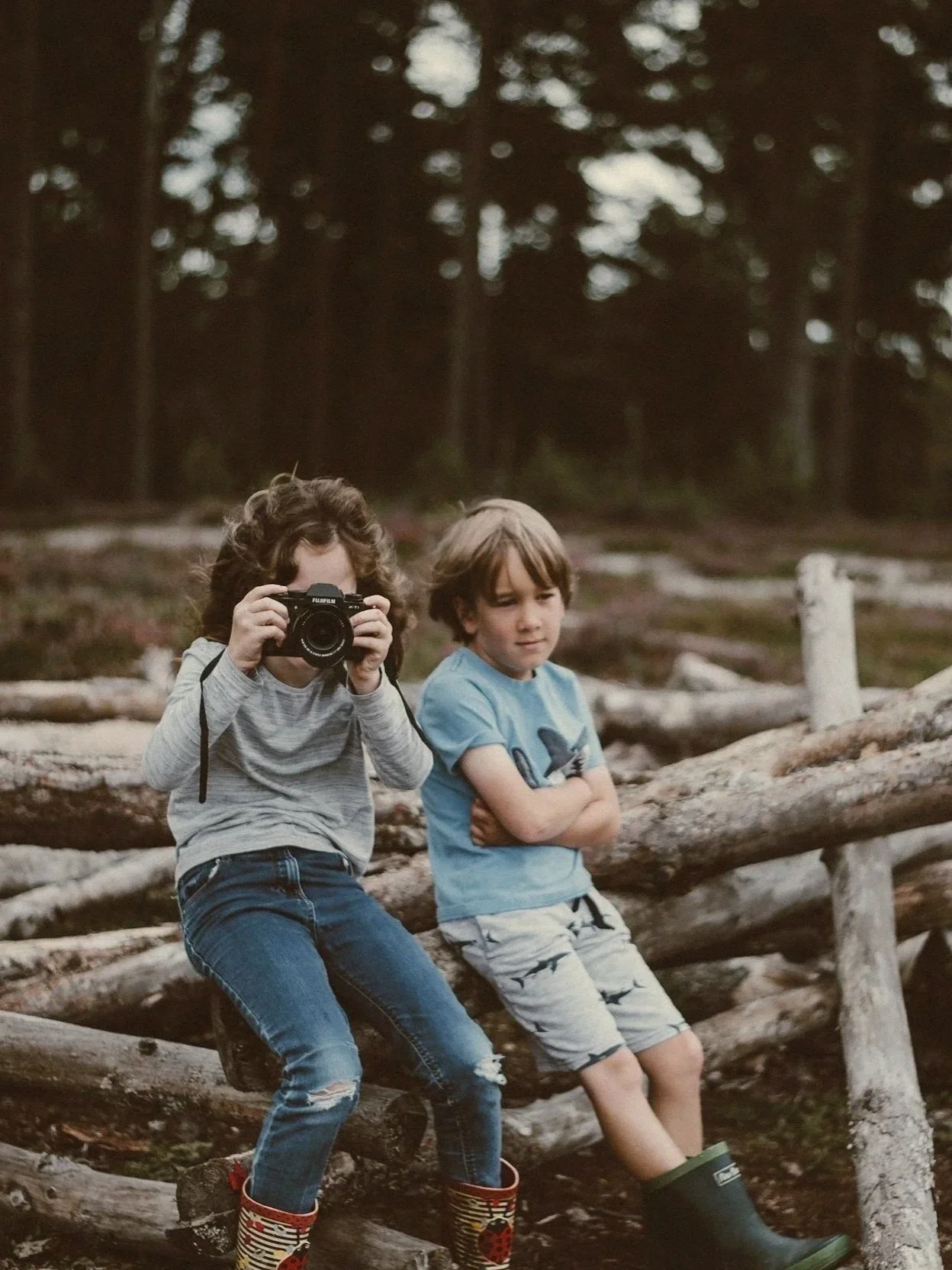 Two children sitting on logs in a wooded area, one girl holding a camera and taking a picture, the boy sitting next to her with arms crossed, both looking thoughtful.
