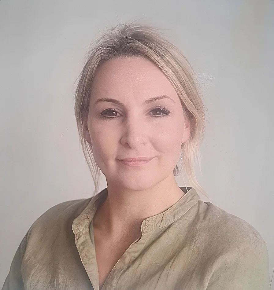 A woman with short blonde hair and light makeup, wearing a beige blouse, standing in front of a plain light-colored background.