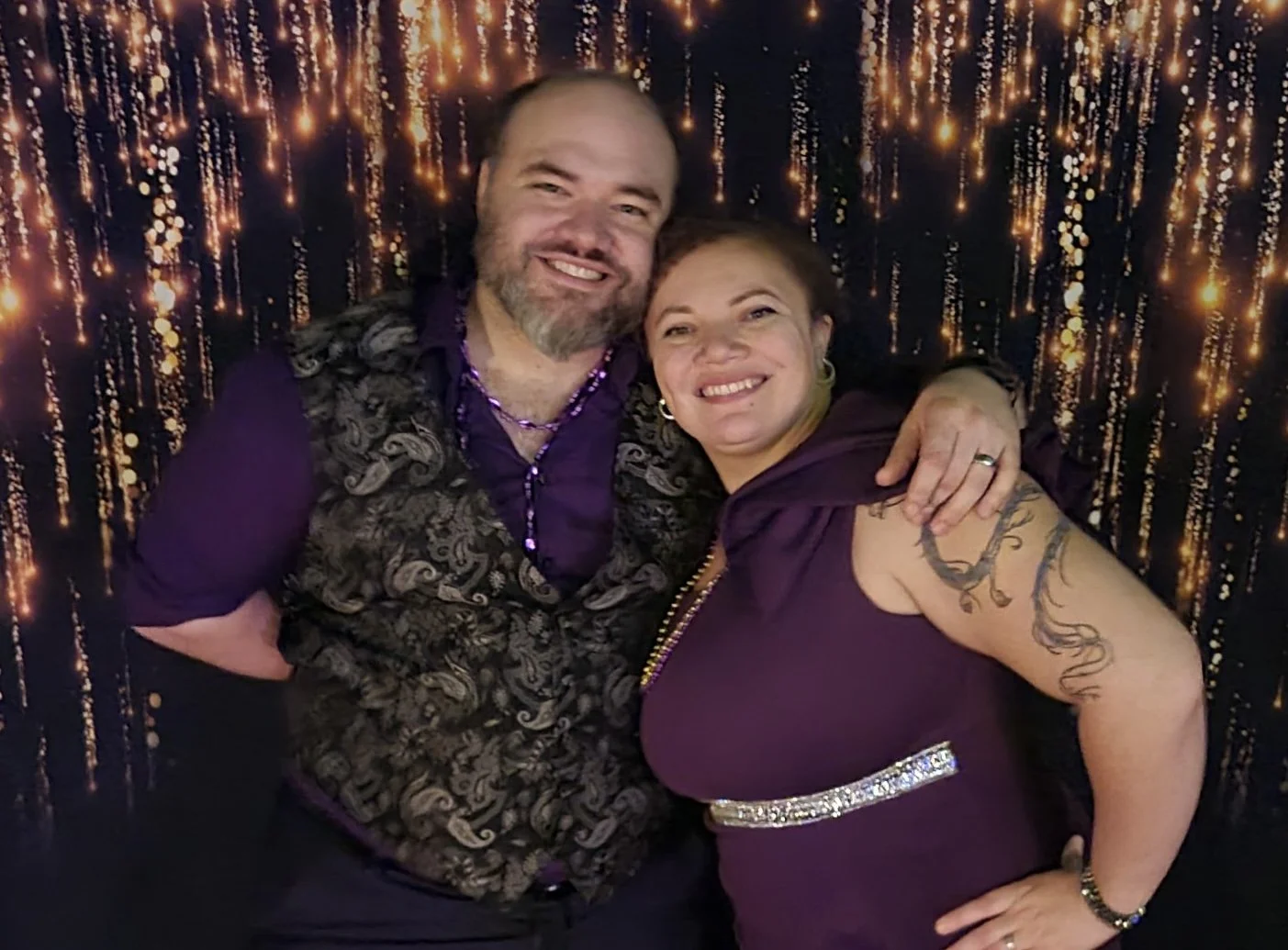 A smiling man and woman embrace each other against a backdrop with hanging lights. The man has a beard and is wearing a patterned vest and purple shirt, while the woman has tattoos on her arm and is wearing a purple dress with a rhinestone belt.