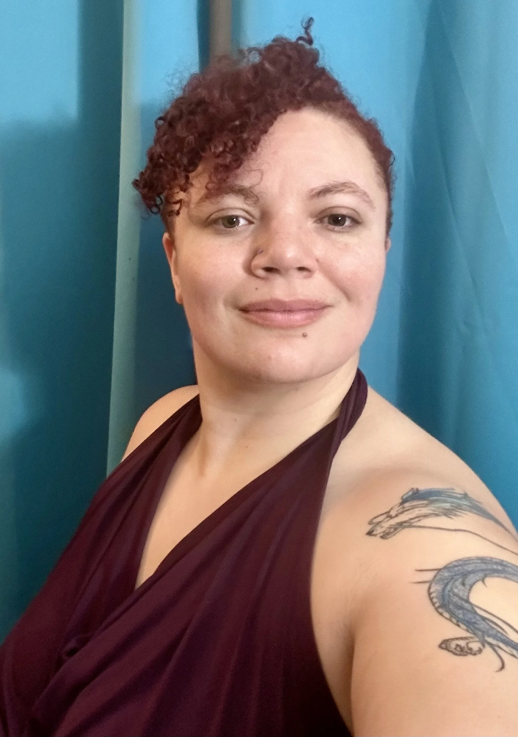 A woman with short curly red hair and a tattoo of a dragon on her arm, wearing a dark purple halter top, standing against a blue curtain background.
