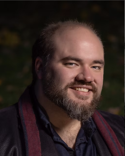 A man with a beard and short hair smiling outdoors in a dark, blurred background.