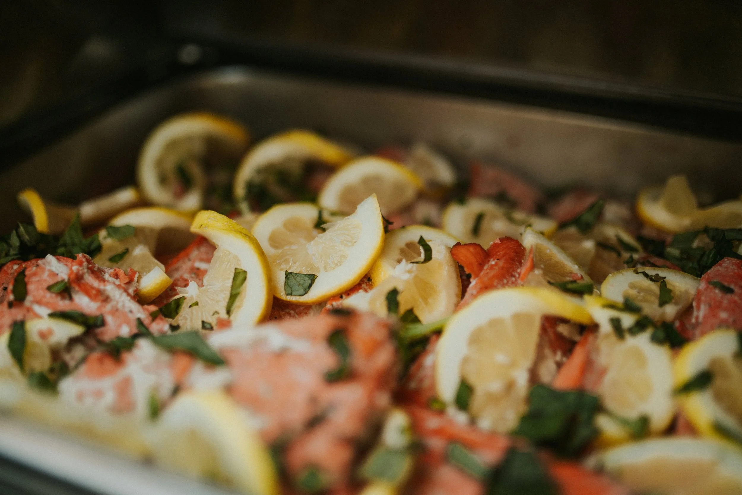 A close-up of salmon fillets topped with lemon slices and fresh chopped green herbs in a silver tray.