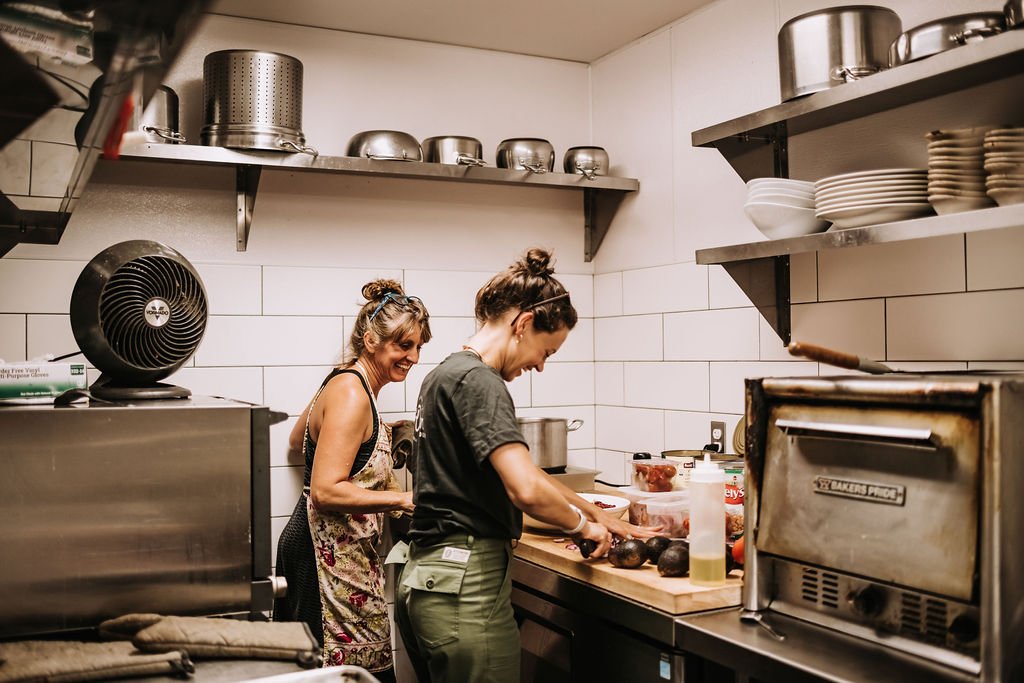 Two women smiling while chopping veggies in a kitchen. There are stainless steel shelves with small appliances, mixing bowls, and dishes in the background.