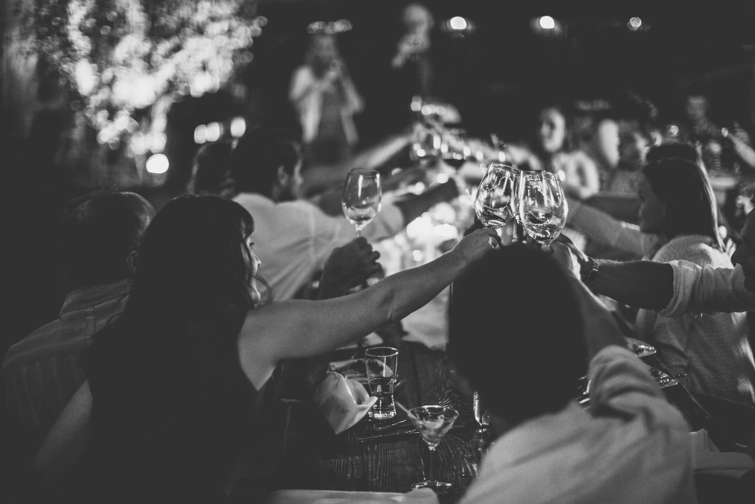 A black and white, lower-contrast image of a large group of people in semi-formal attire toasting wine glasses at a table outside.