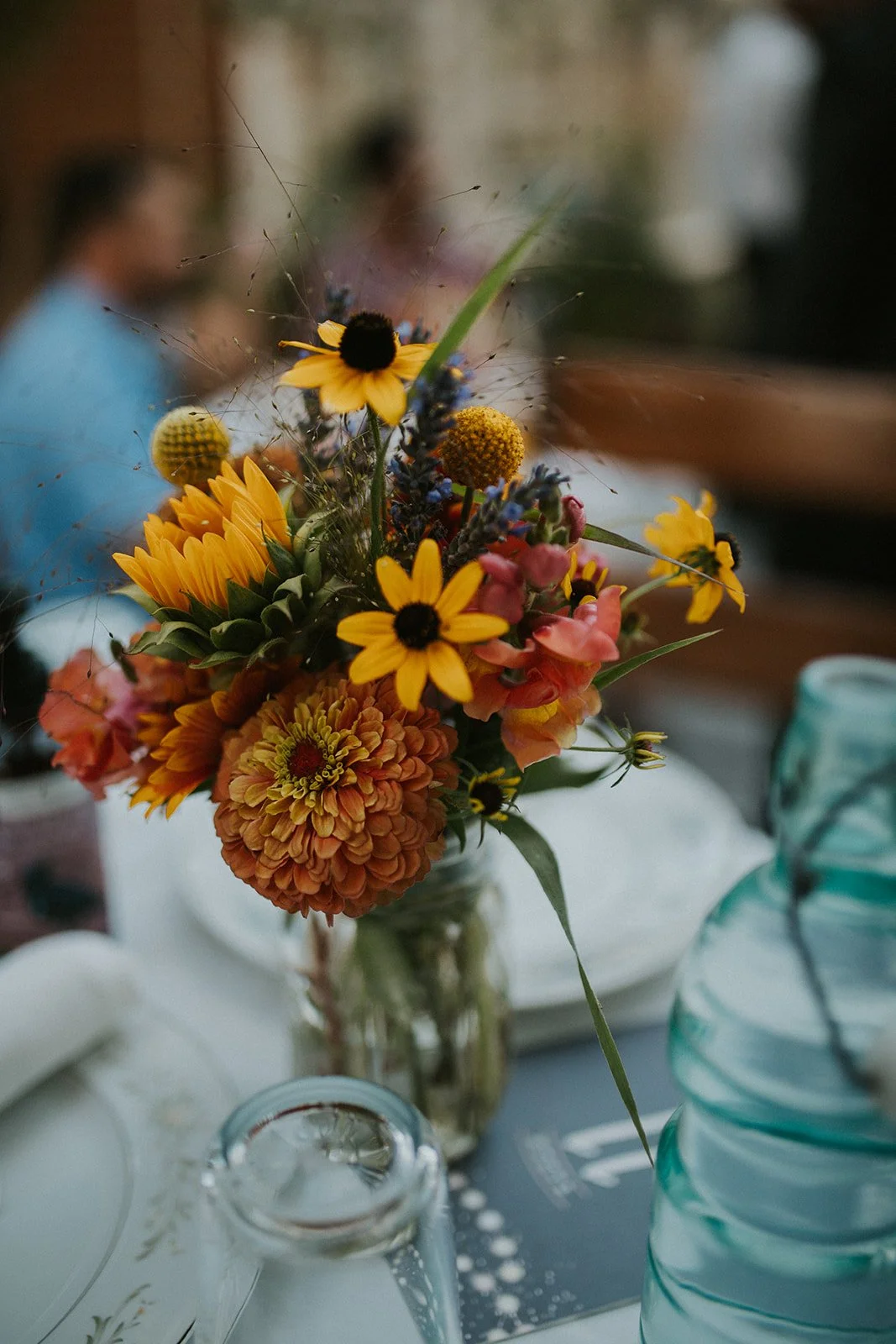 A close-up on a vase of flowers set on a table as part of a place setting. It contains flowers in shades of orange, pink, red, and yellow.