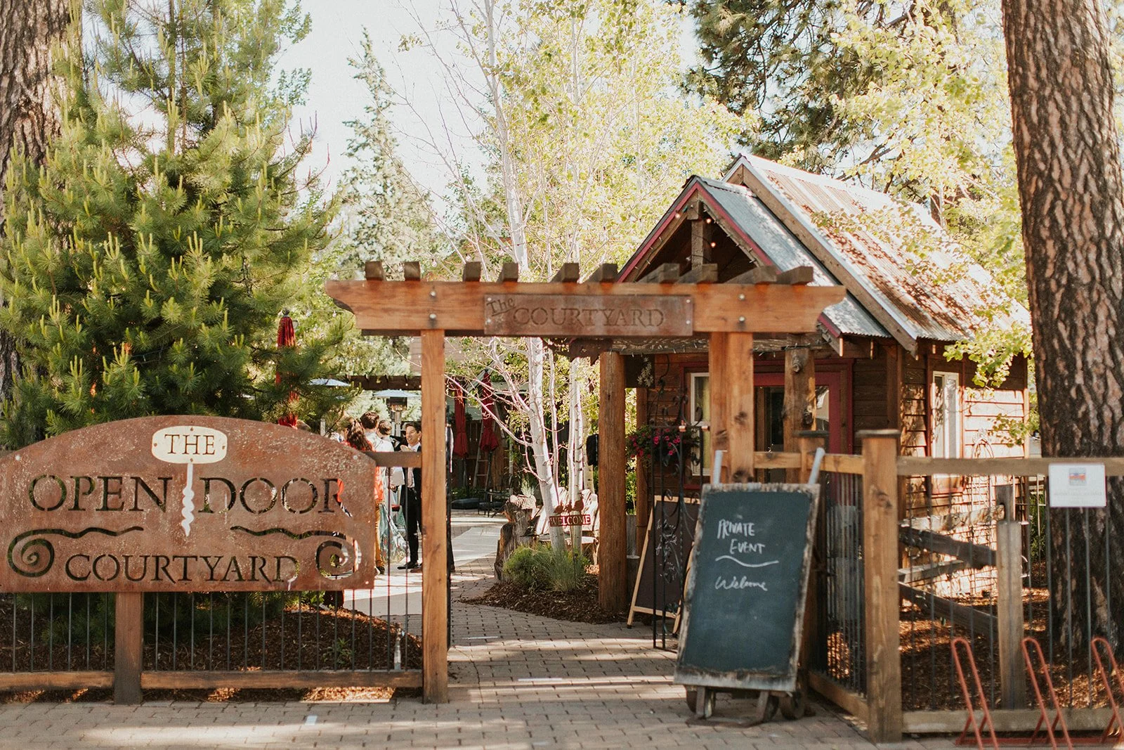 The exterior of The Open Door outdoor courtyard. There is a wooden fence and trellis in the foreground with a sign reading "The Open Door Courtyard". In the background, there is a winding path with smaller buildings and trees.