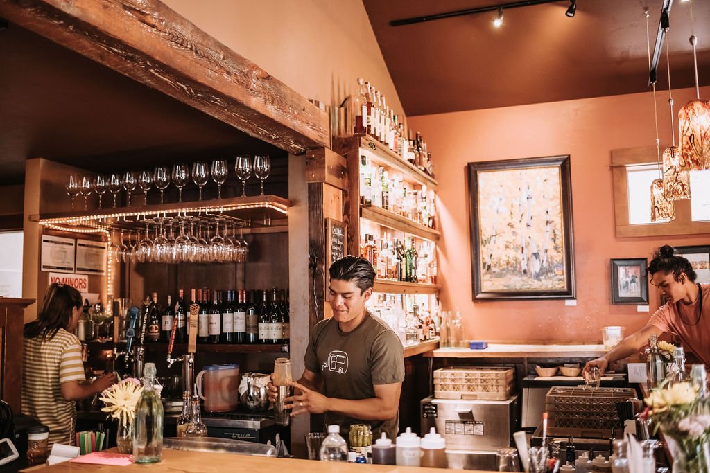 A male bartender in a brown shirt shakes a cocktail behind a wooden bar. Shelves with bottles and wine glasses are visible in the background.
