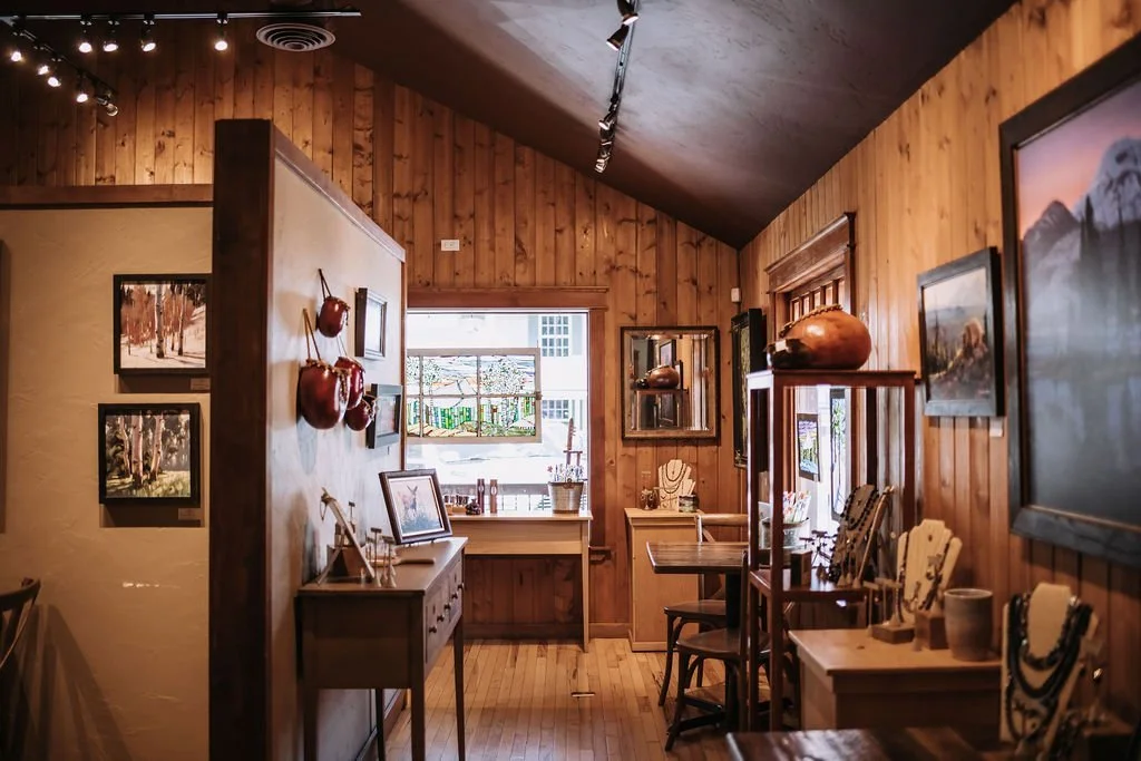The interior of an art gallery with wood-paneled walls displaying artwork and furniture, with a window in the background.
