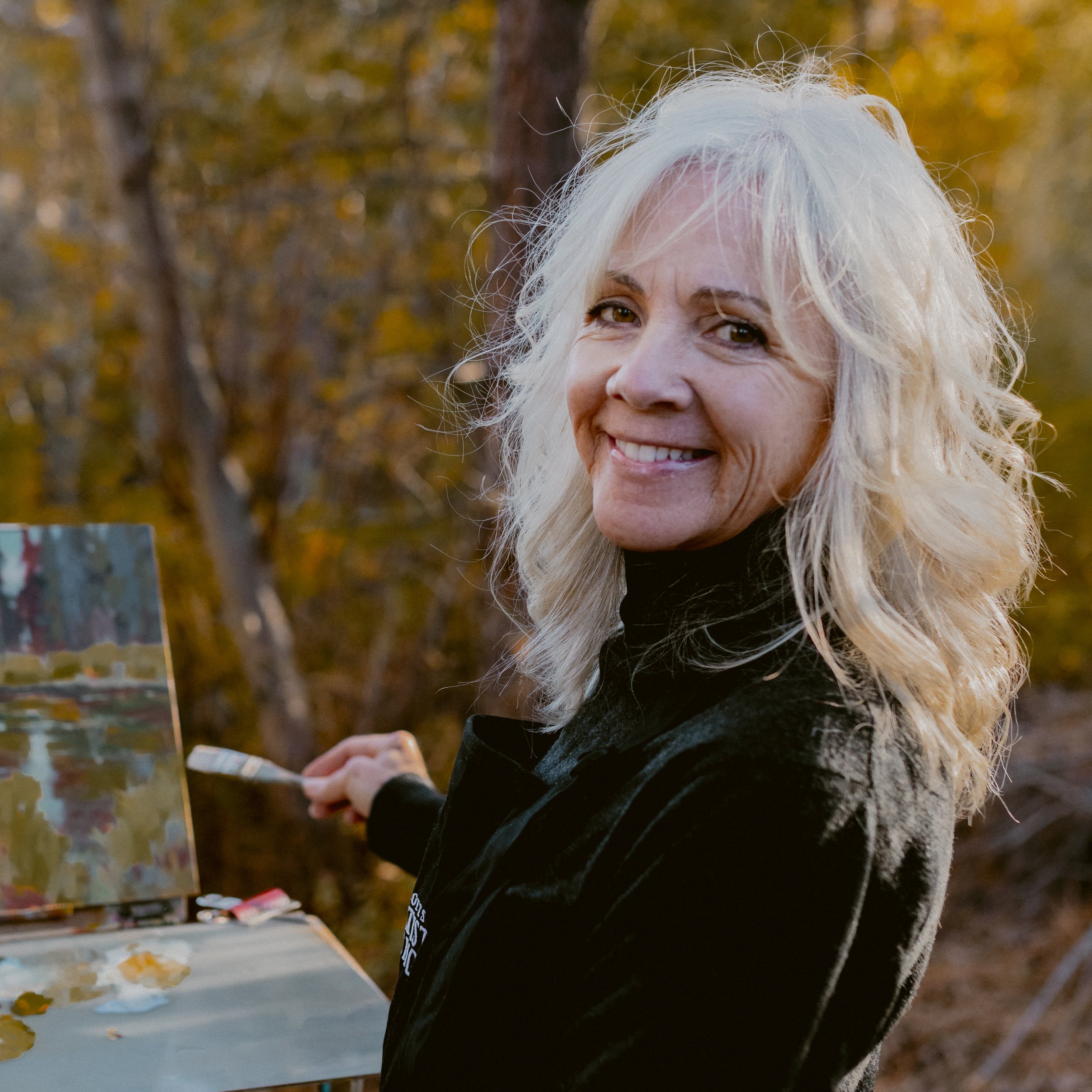 Photo of Sarah B Hansen turned towards the camera while smiling, while holding a paintbrush in her right hand. An easel with a painting is in the background.