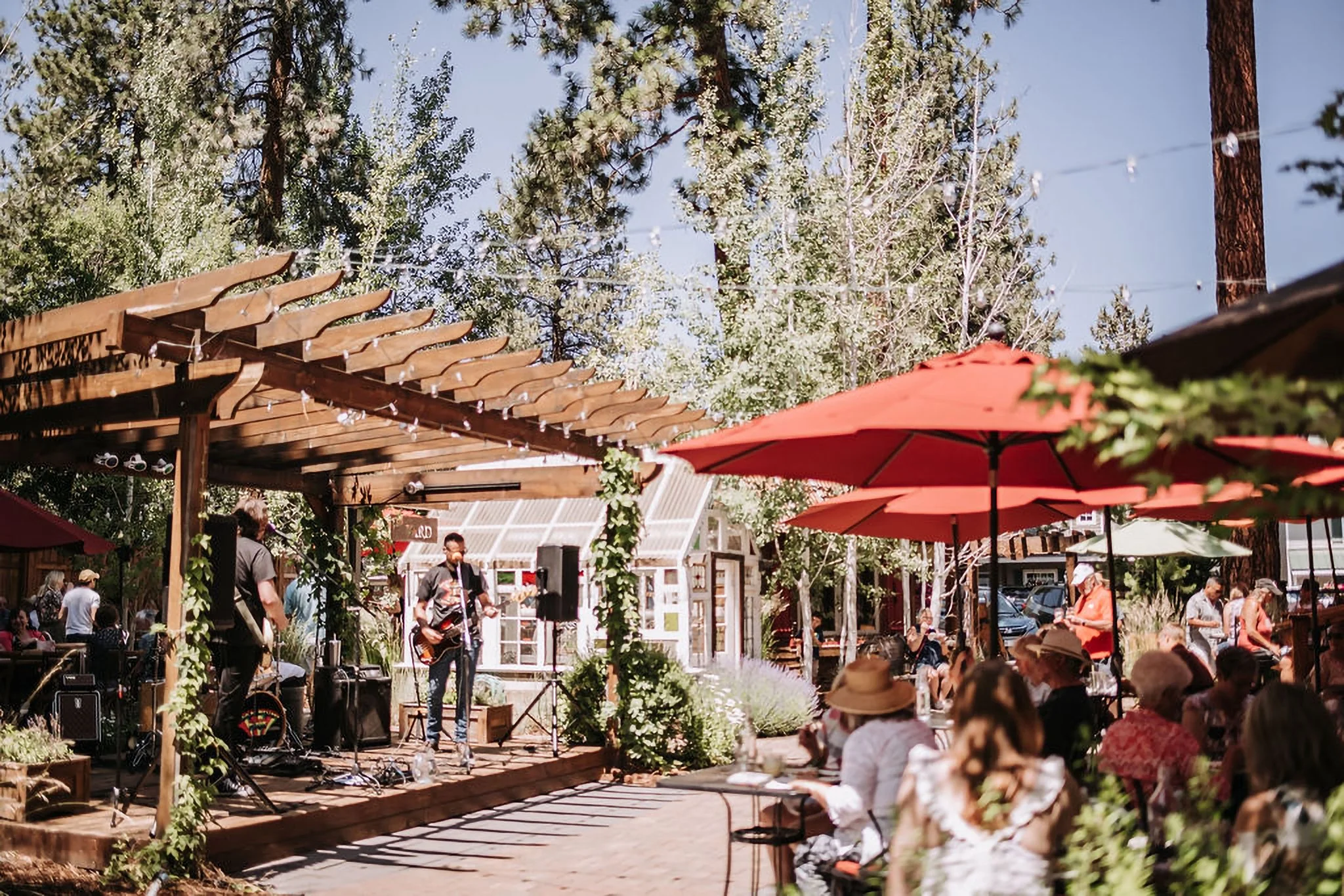 A small group of musicians perform on an outdoor wooden stage on the left of the frame, while tables with diners under red umbrellas watch from the right. There are trees and a blue sky in the background.