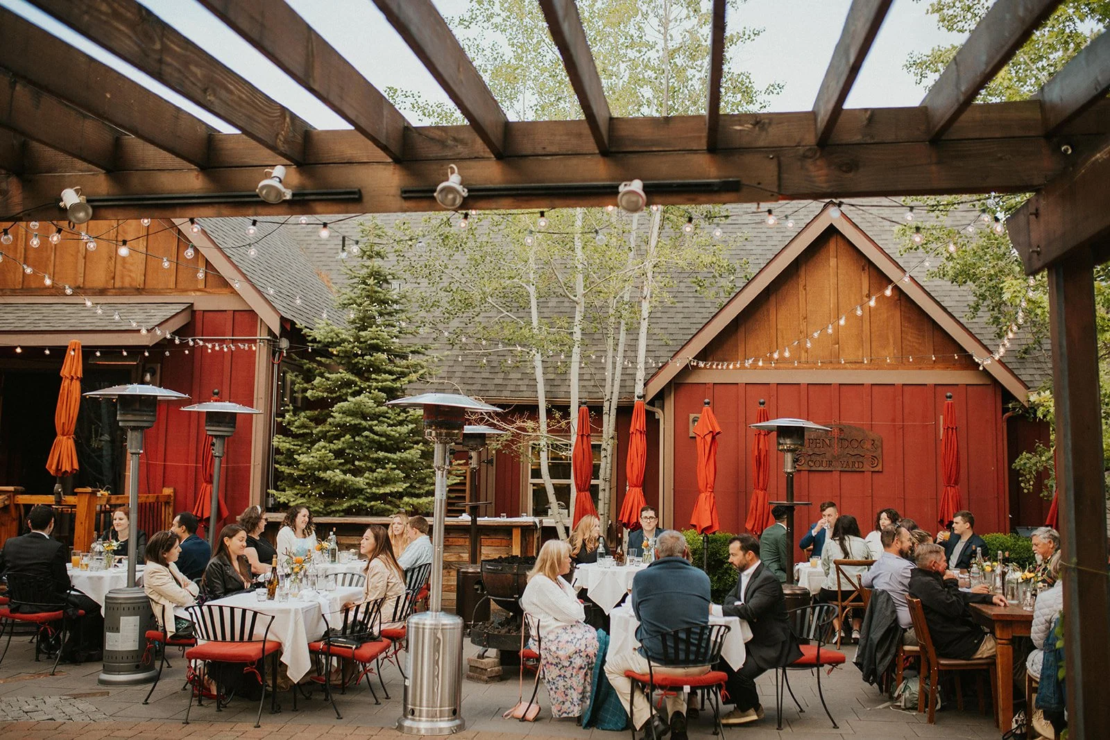Looking out from under a wooden trellis at groups of people seated around tables with white tablecloths. They're in an outdoor courtyard with string lights above and a barn red building with trees behind.