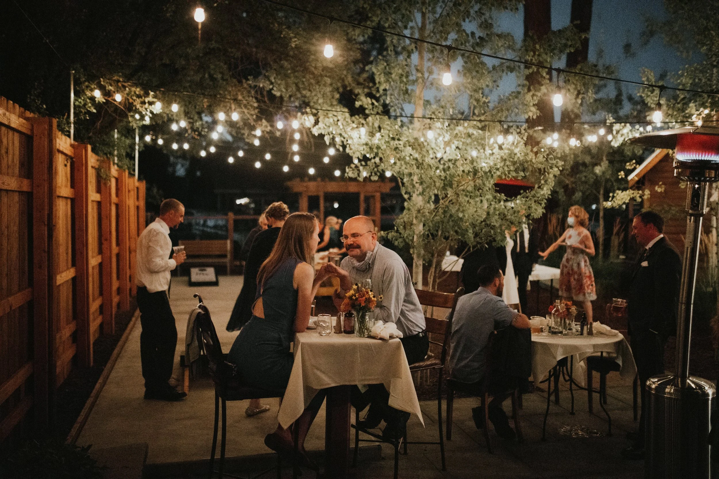 A couple is sitting at a table in a courtyard outside, at night, under illuminated string lights. There are other couples are seated at different tables behind them, with trees in the background.