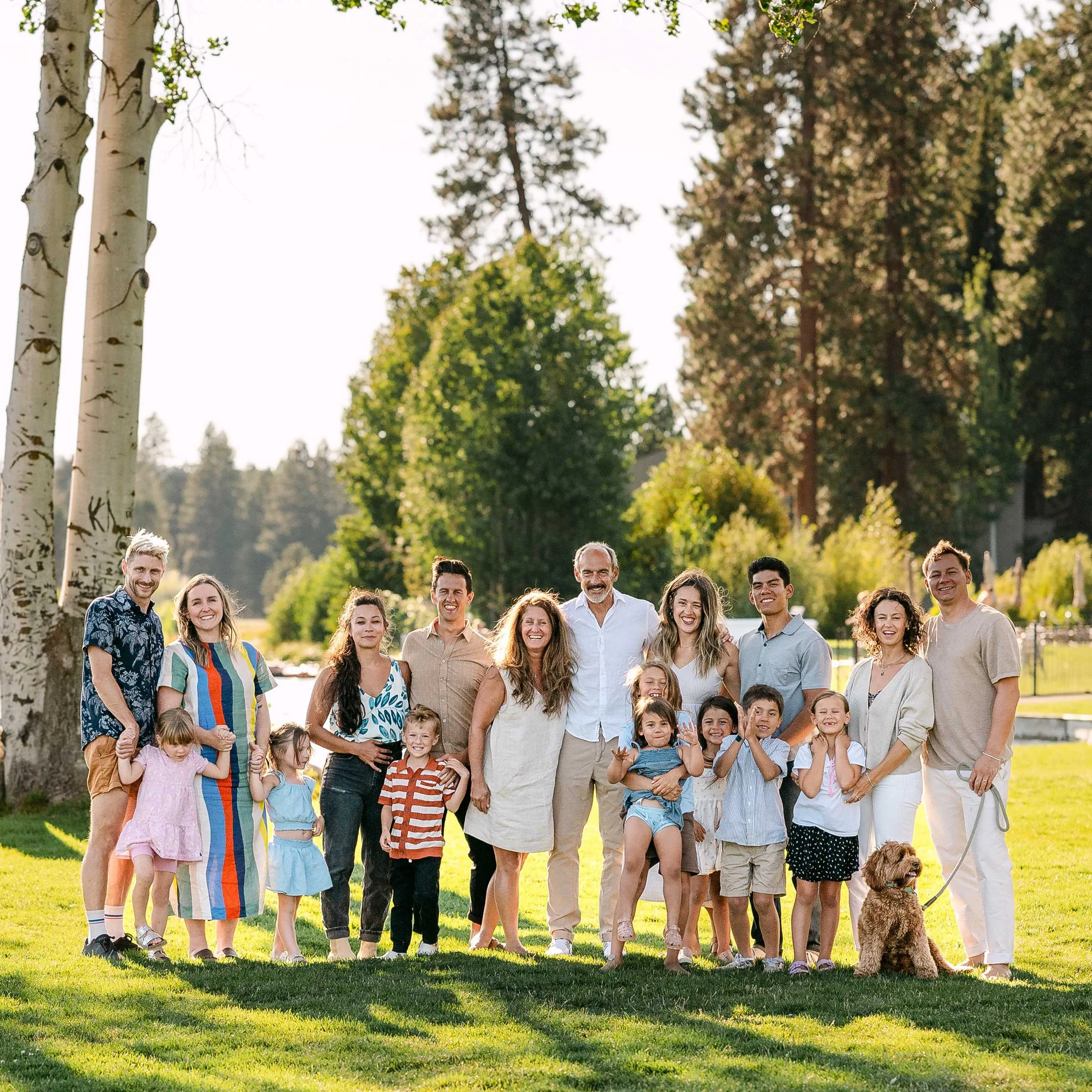A group of smiling people standing in a green, grassy field with soft backlighting and trees behind them.