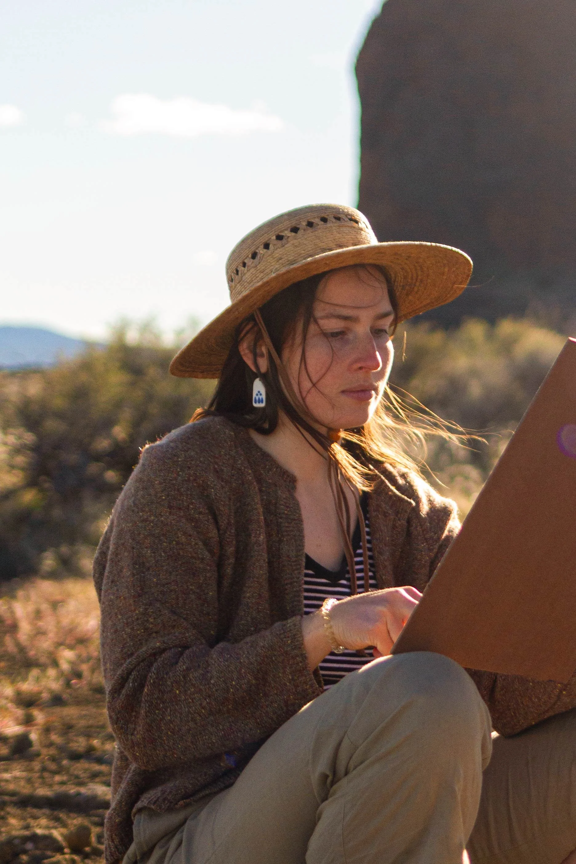 A photo of Taylor Manoles looking down at a board. The sun is coming from the upper-right edge, and she is sitting against a high desert nature background