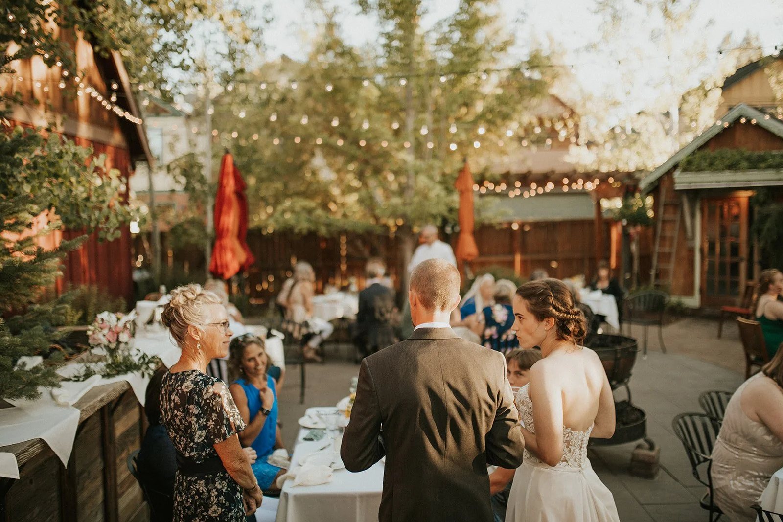A bride and groom face away from the camera towards a table in an outdoor courtyard with people seated around it, chatting. There are other tables in the courtyard behind them, with string lights above and a wooden fence and trees in the background.