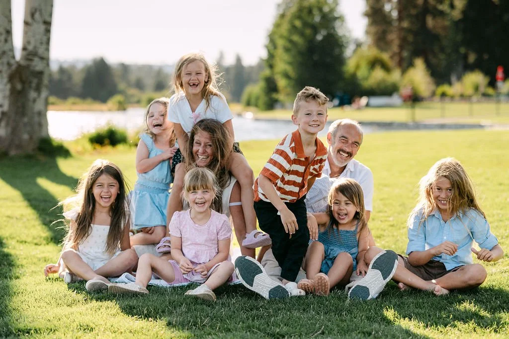 A group of laughing kids sitting on the grass with two adults smiling at the camera. The image is softly backlit, with water and trees in the background.