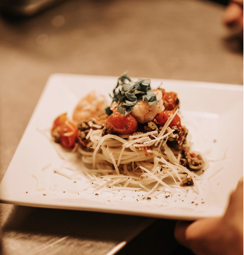 A close-up of angel hair pasta on a plate, topped with cherry tomatoes, shredded cheese, and chopped fresh herbs.