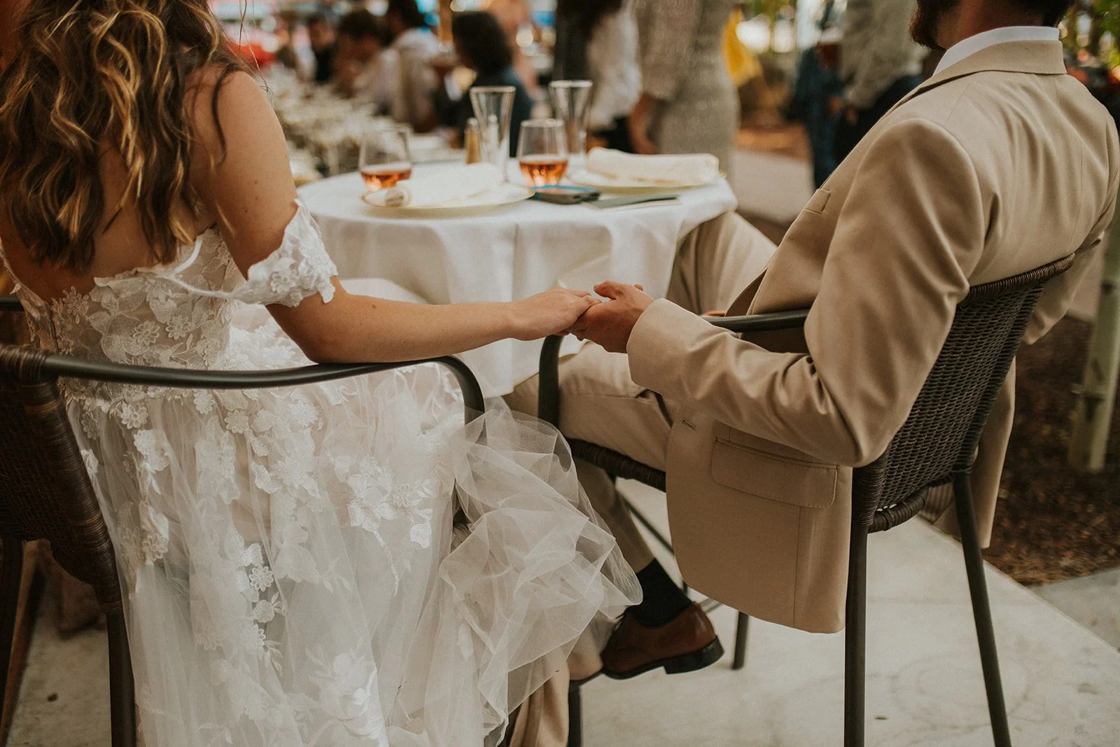 A close-up of a woman in a white wedding dress and a man in a tan suit seated at a table while holding hands. The image is taken from behind them, with their faces out of frame.