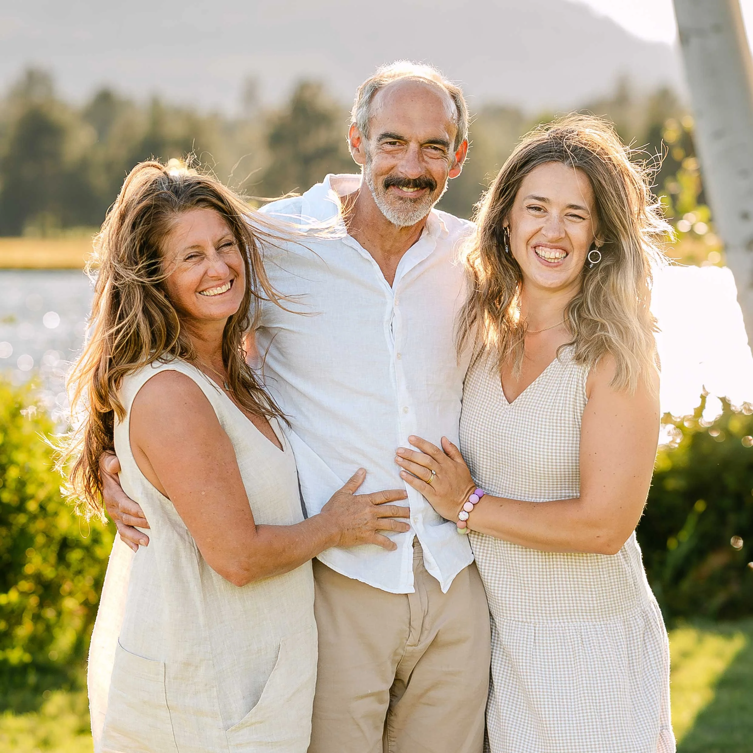 Three people are smiling and facing the camera in light tan clothing, with a green, grassy backdrop and soft backlighting.