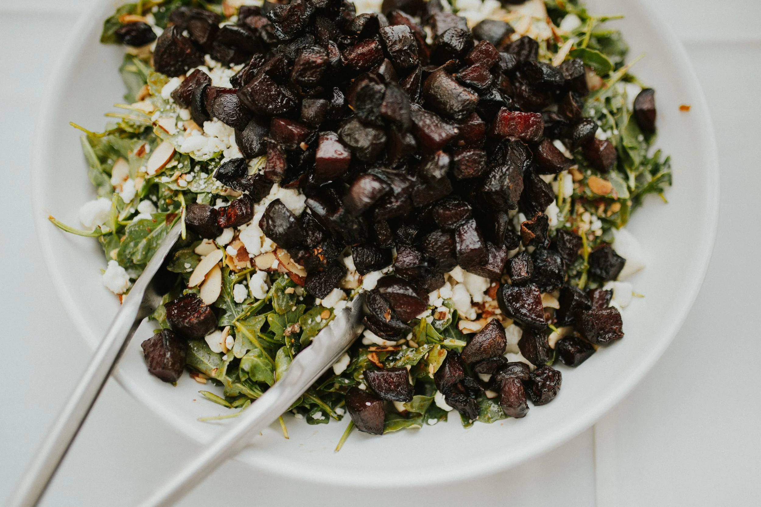 A close-up of silver tongs in a salad bowl filled with bright green arugula, cheese, slivered nuts, and deep red cubed veggies.