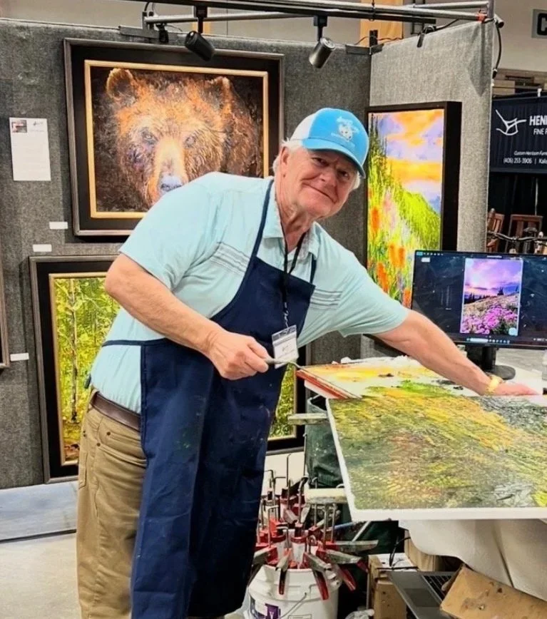 A man in a blue apron, Garth Williams, is smiling at the camera while leaning over a table with a painting laying on it. In the background are paintings hung on portable gallery walls, including one of a bear and others depicting outdoor scenery.