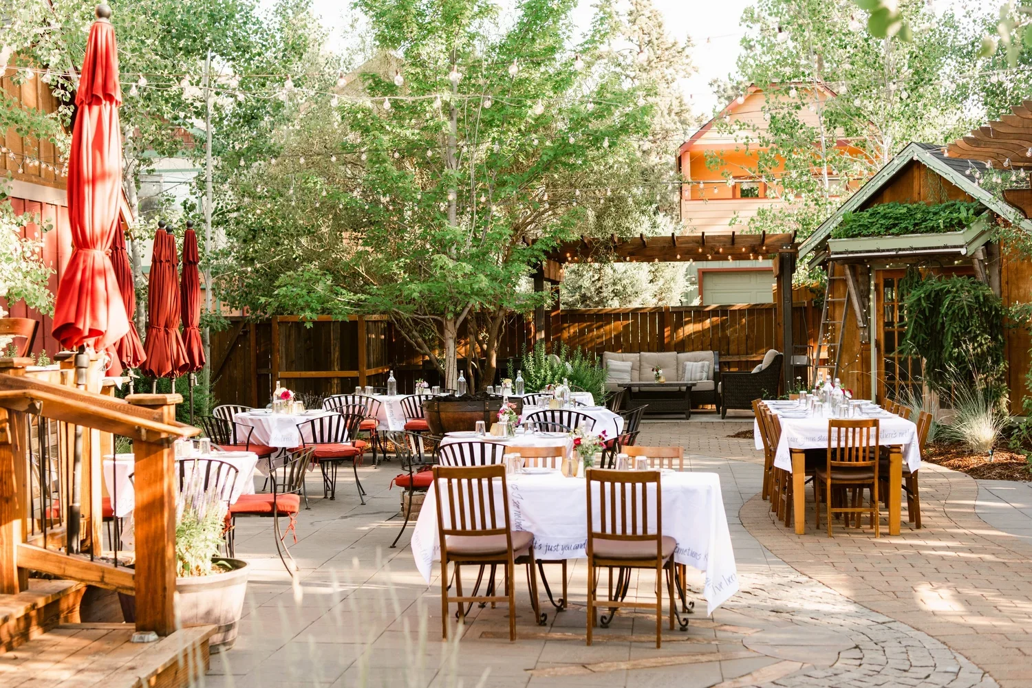 Tables with white tablecloths are arranged in an outdoor courtyard with string lights above them. There are wooden buildings, trees, and a wooden fence in the background.
